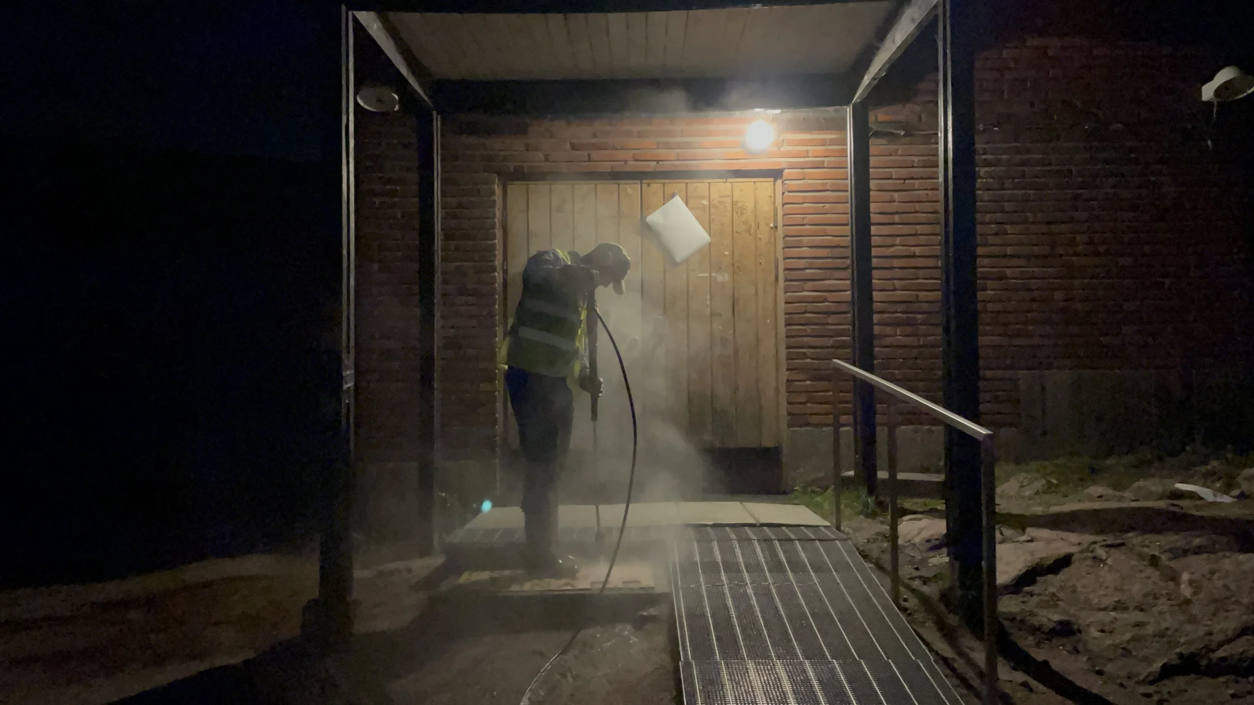 Person pressure washing a surface at night under a light, near a wooden door and brick wall.