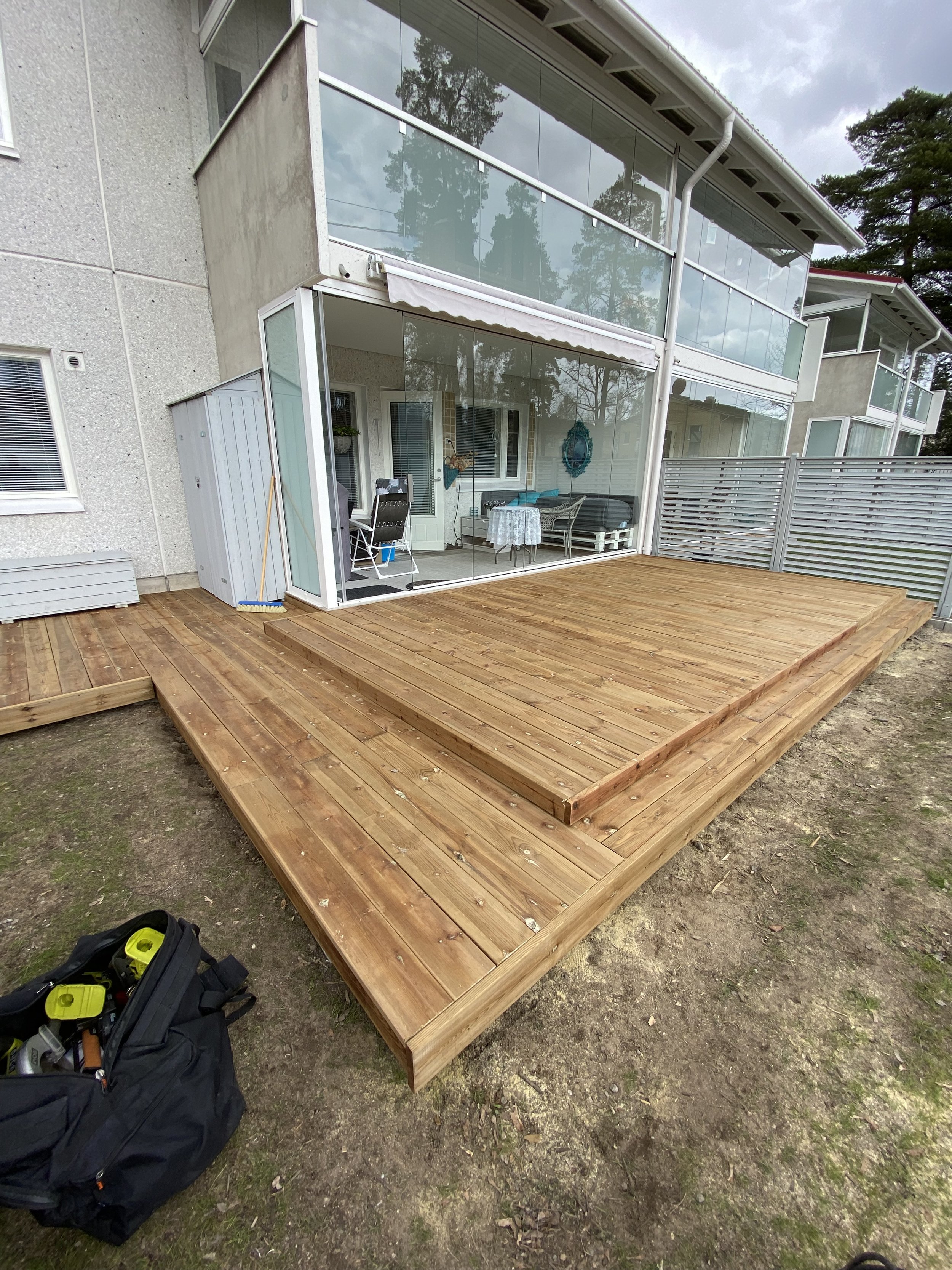 A wooden deck attached to a modern house with glass railings, outdoor furniture, and a bag of tools on the ground.