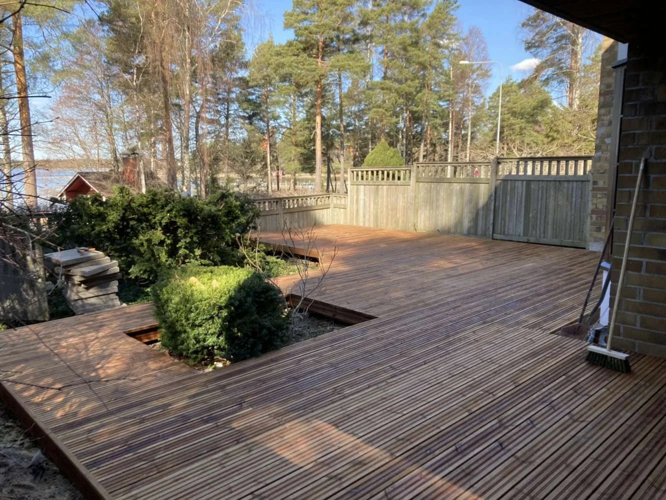 Wooden deck with square cutouts featuring shrubs, surrounded by a wooden fence and trees, under a blue sky.