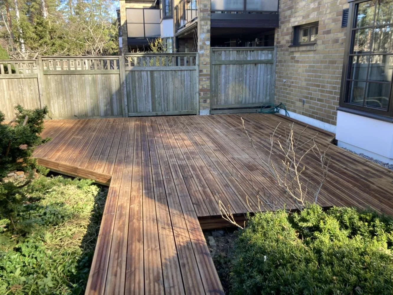 Wooden deck in a backyard with greenery, next to a brick house and wooden fence.