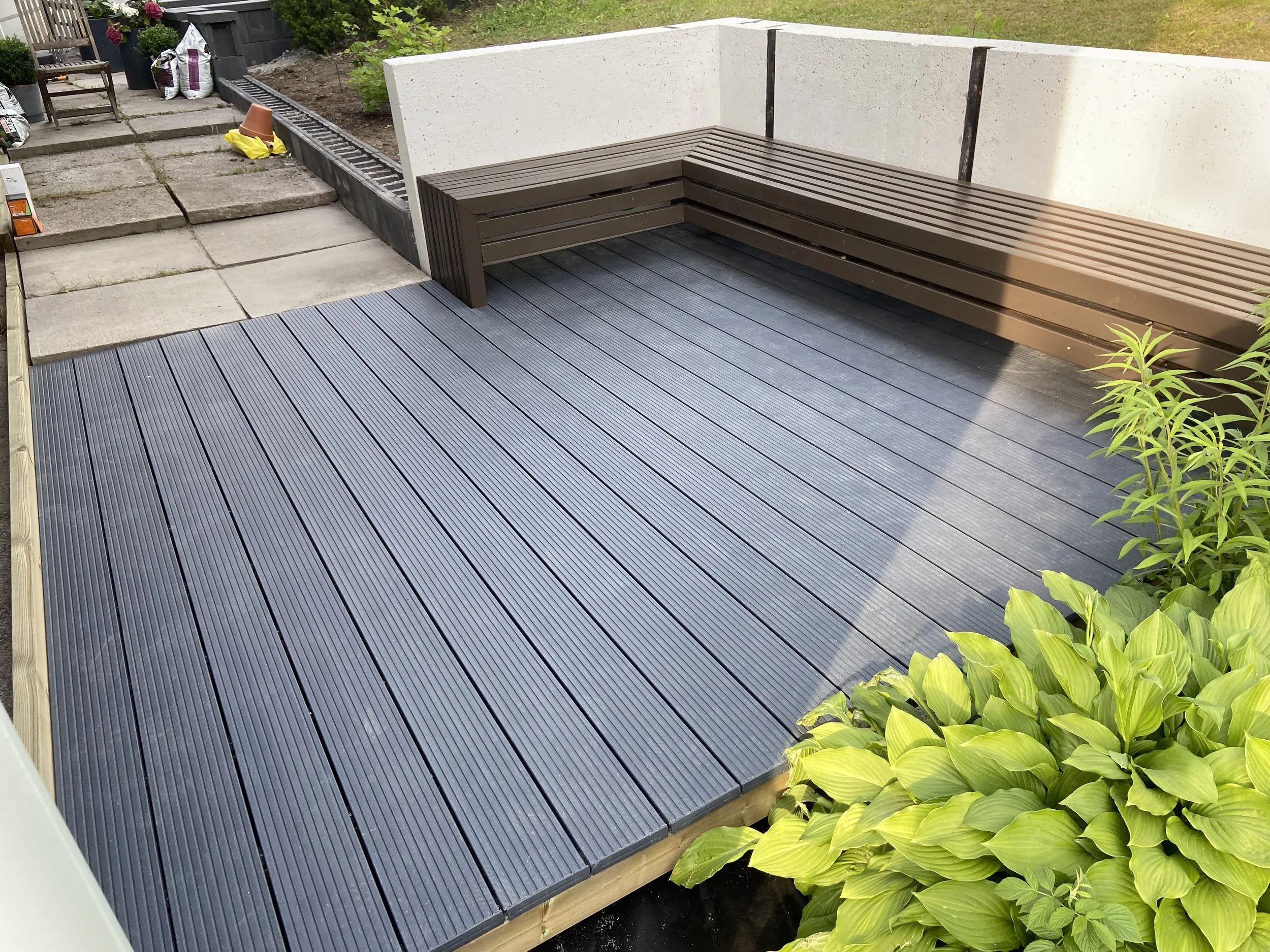 Gray composite deck flooring with a wooden bench along a white wall, surrounded by green plants.