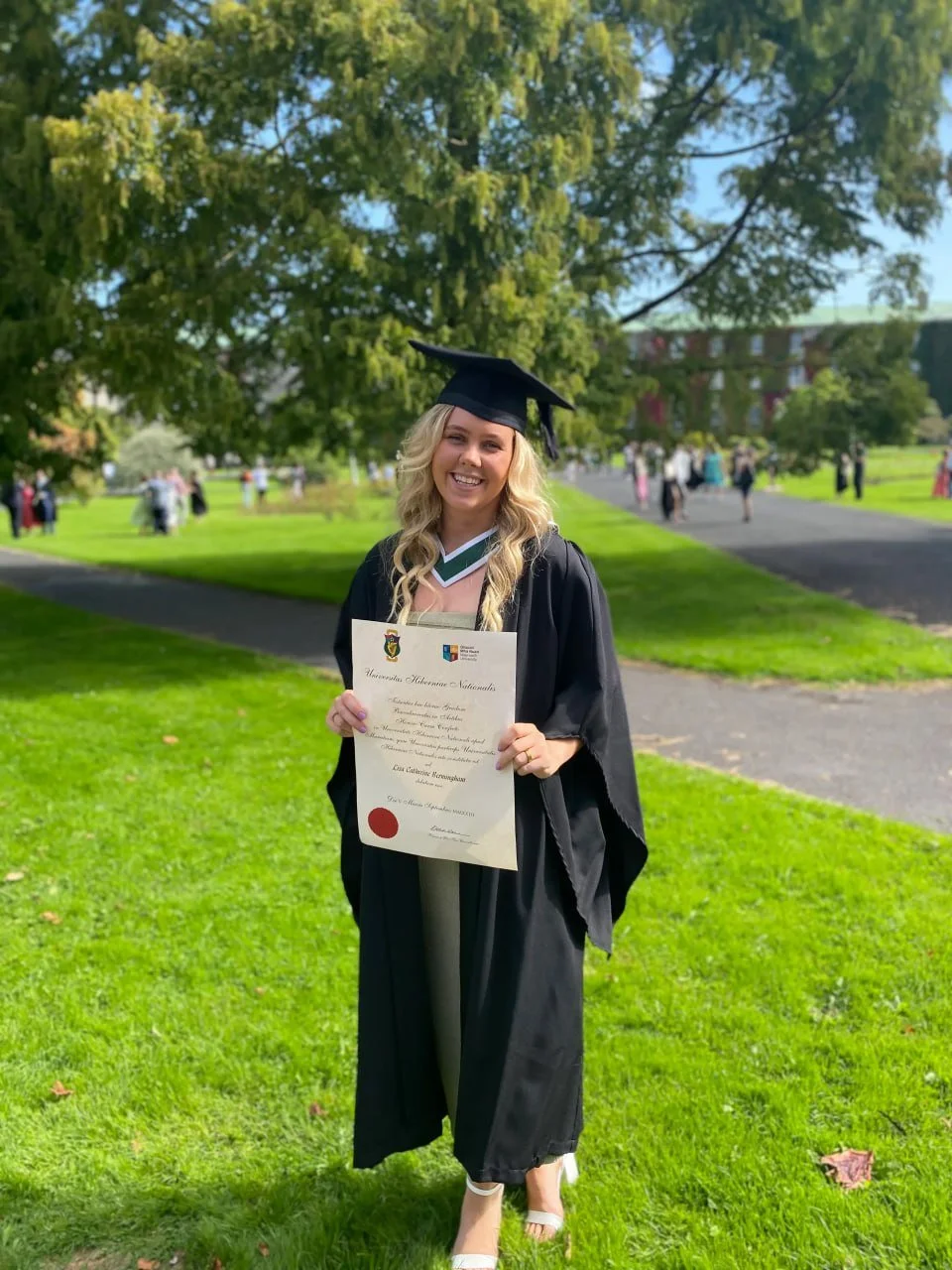 A young woman in a graduation cap and gown stands outdoors on a green lawn, holding a diploma and smiling at the camera, with trees and other graduates in the background.