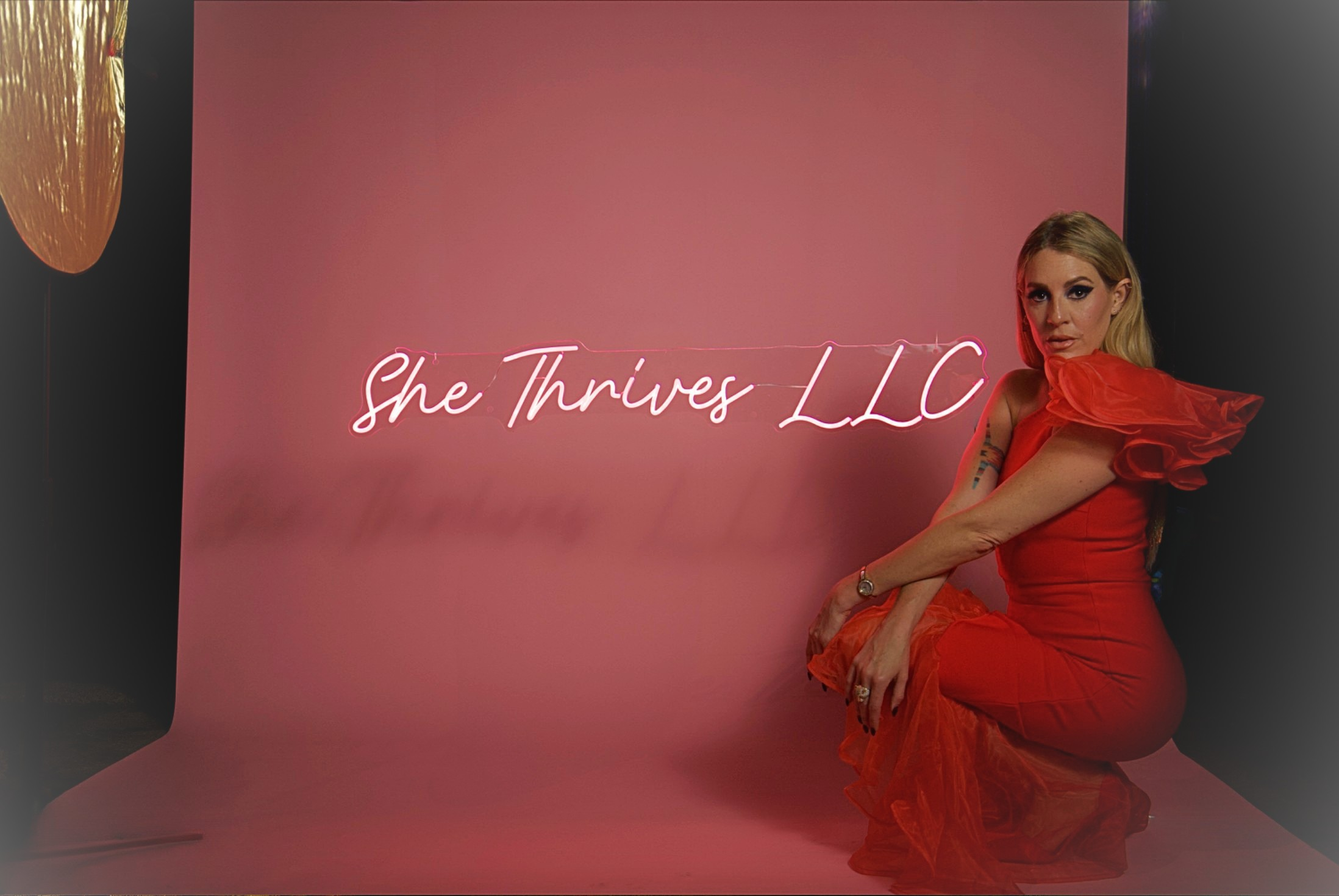 A woman in a red dress posing in front of a pink backdrop with a neon sign that reads 'She Thrives LLC'.