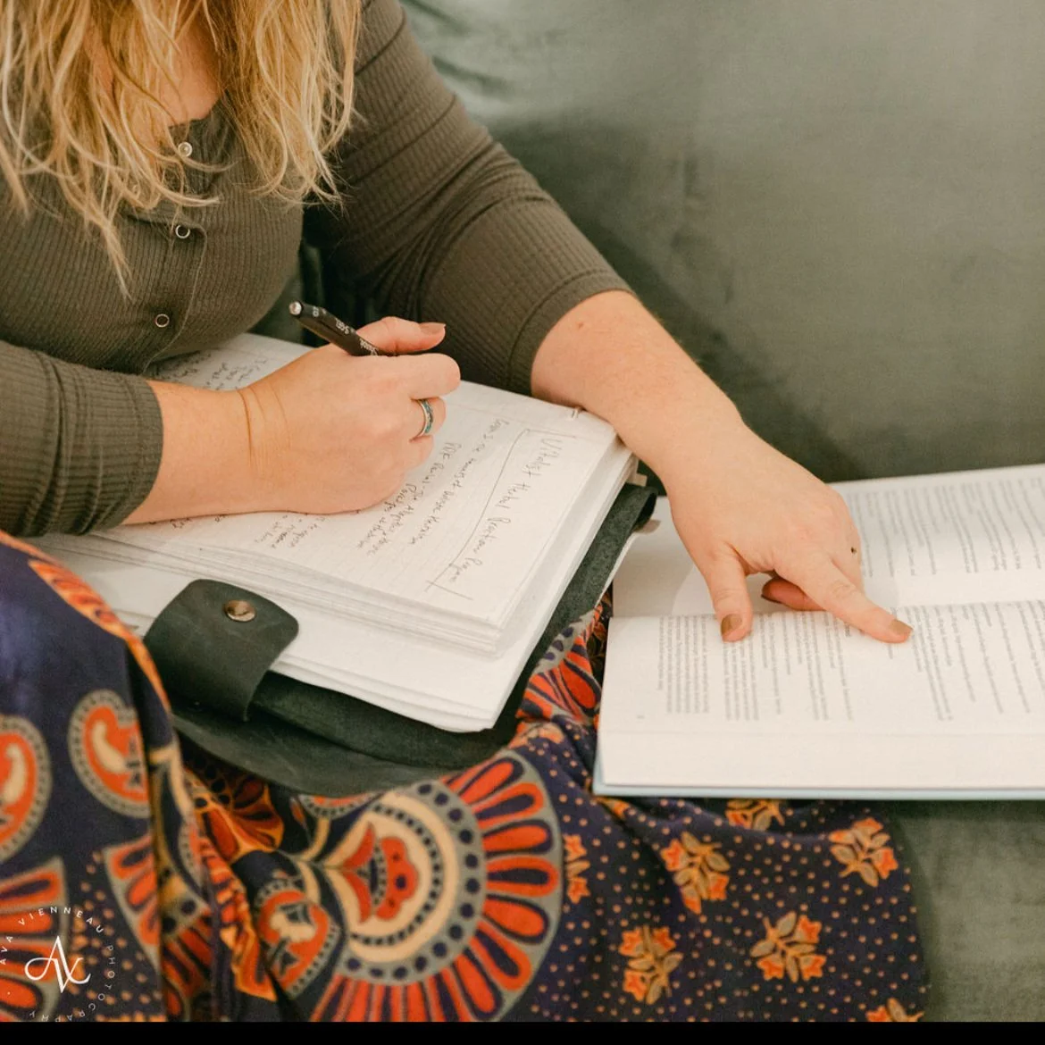 A person with long curly hair taking notes in a notebook with handwritten notes and a pie chart, while pointing to a printed book or document with their right hand. They are wearing a dark gray ribbed long-sleeve shirt and a patterned skirt with orange, yellow, and navy blue designs.