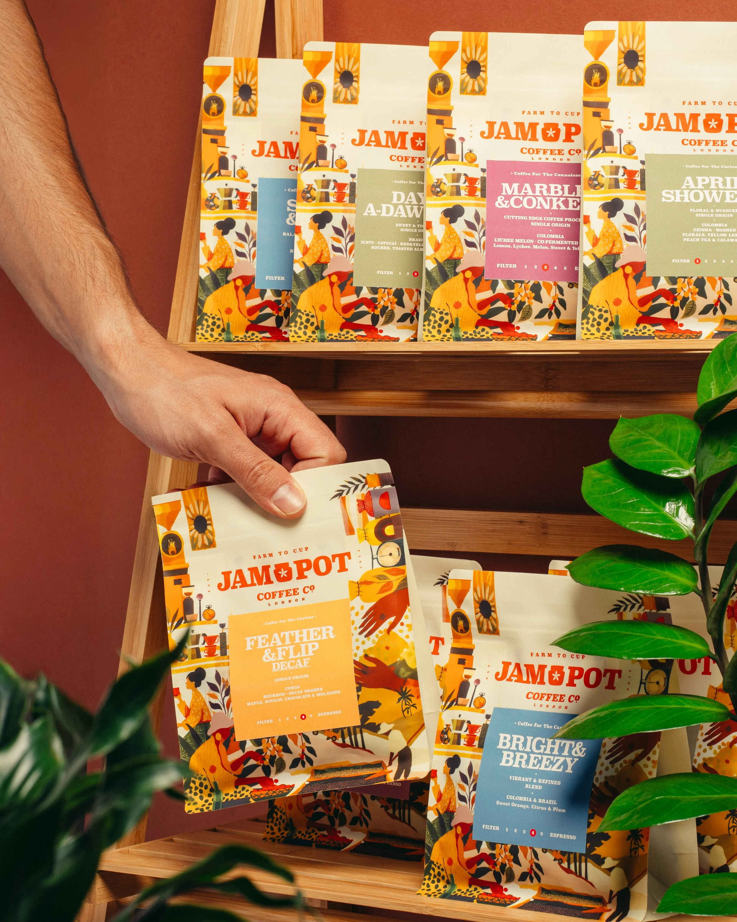 Hand removing a bag of Jampot Coffee Co. labeled 'Feather & Flip De Cafe' from a wooden shelf with other colorful coffee bags labeled 'Bright & Breezy' and 'Marble & Cone'.