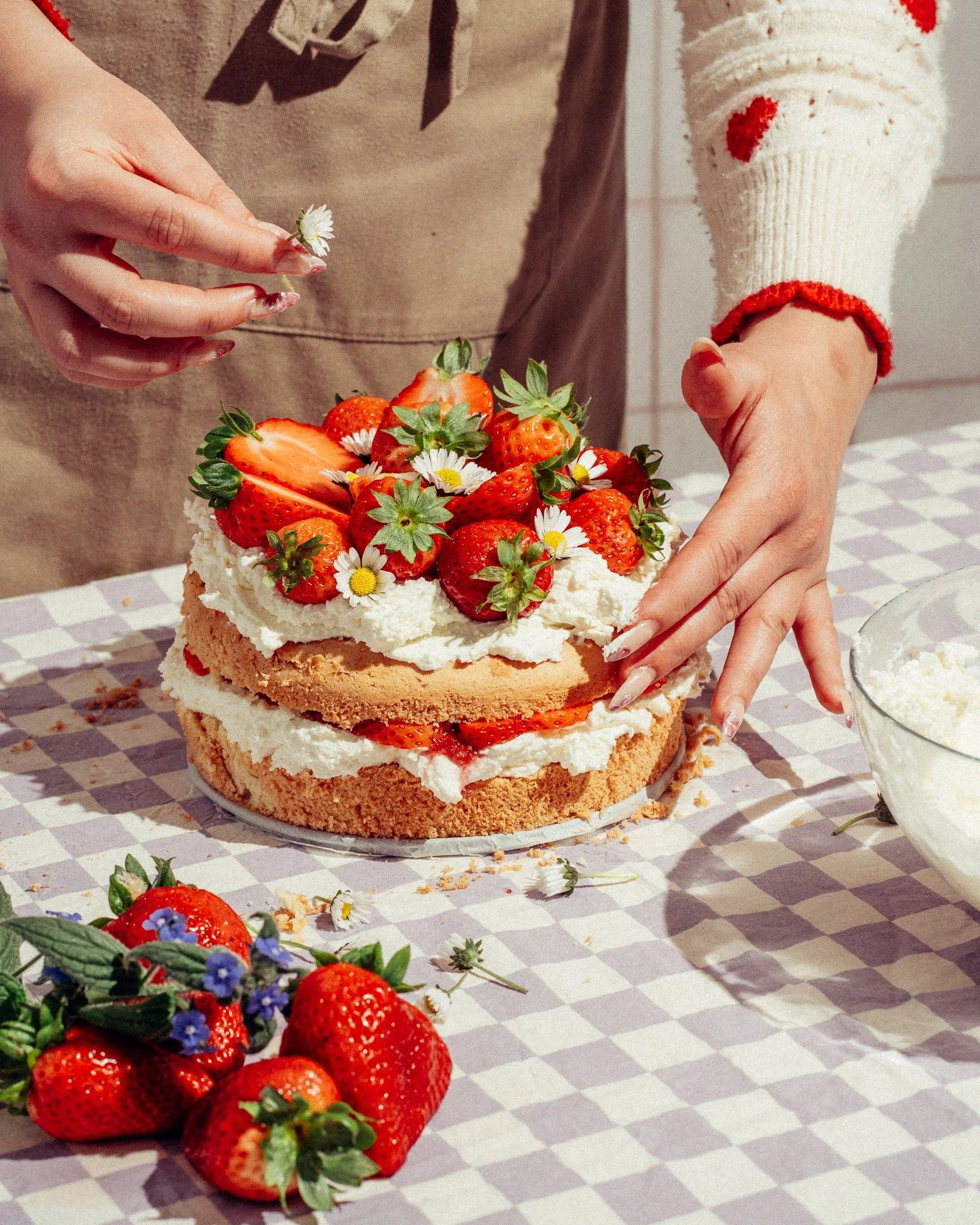 A person decorating a layered strawberry cake with strawberries and small daisy flowers, on a checkered tablecloth, with strawberries and flowers in the foreground.