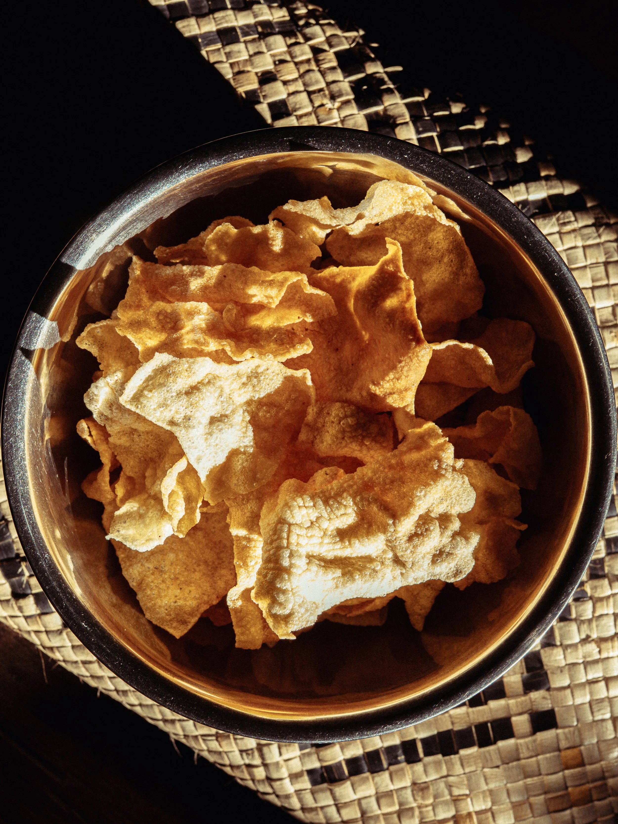 A bowl of potato chips on a woven placemat with a dark background.