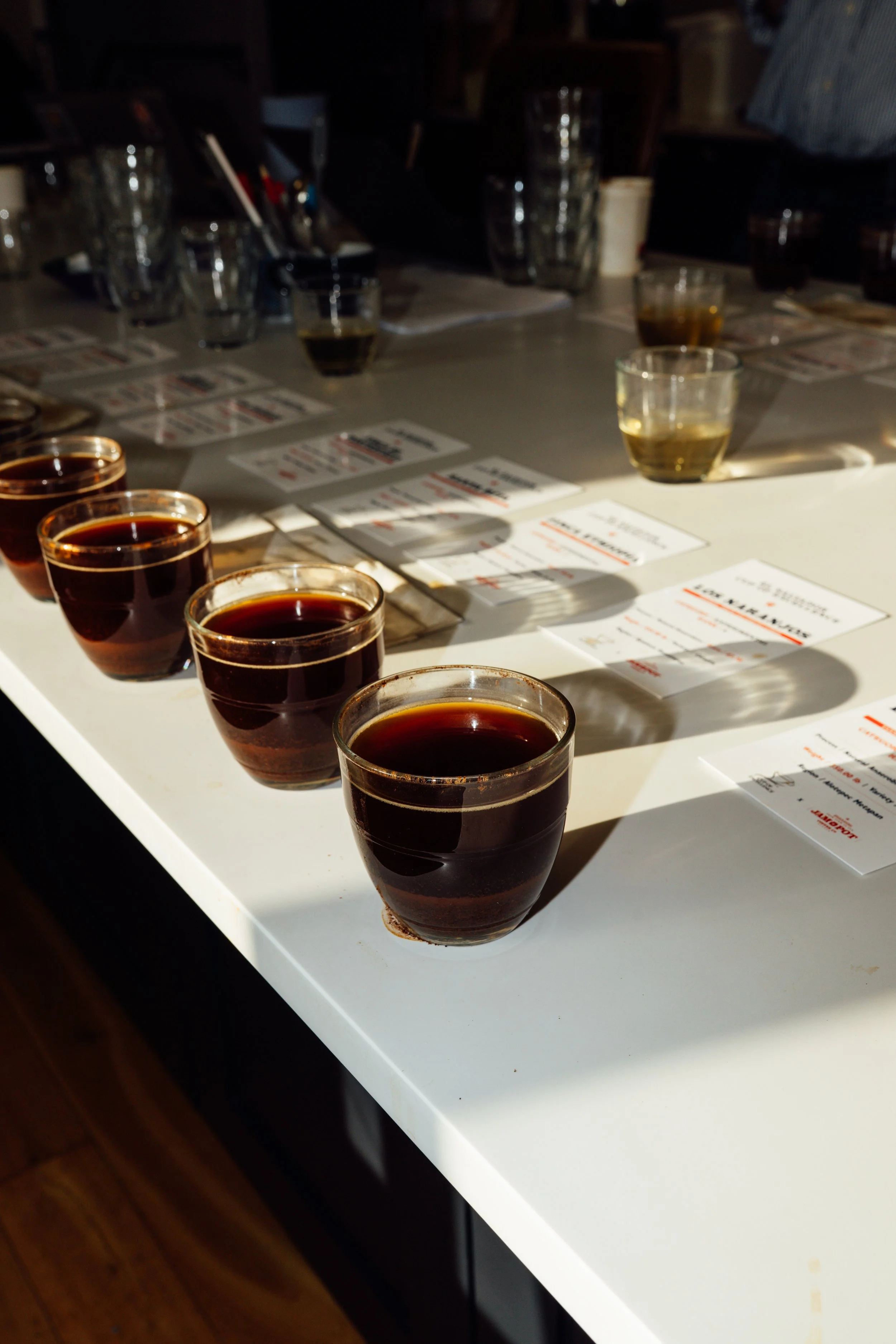 Several small glasses filled with dark liquid, possibly coffee or espresso, arranged on a white table. In the background, there are additional glasses and paper menus.
