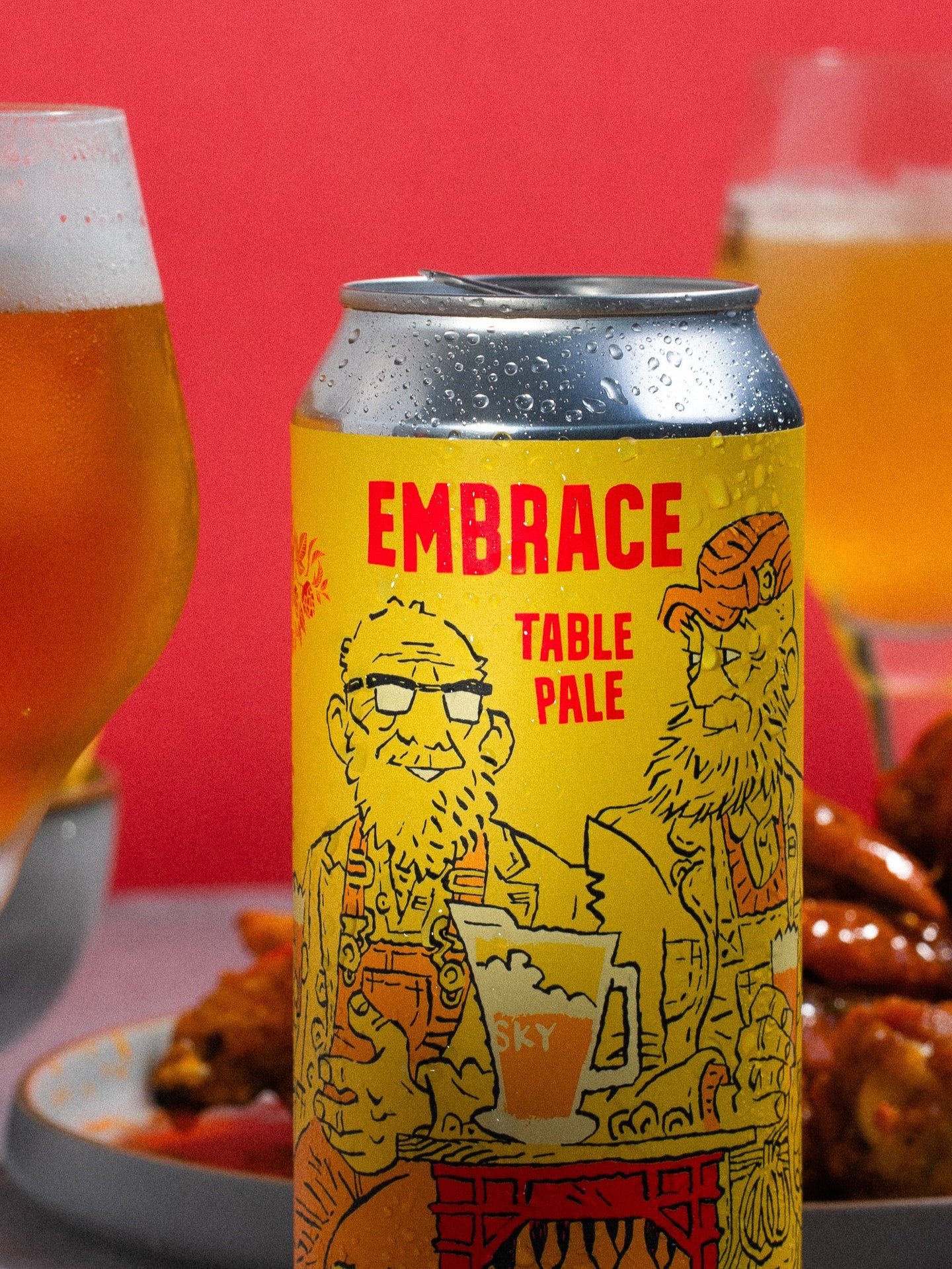 Close-up of a can of Embrace Table Pale beer with artistic design, surrounded by glasses of beer and plates of food, against a red background.
