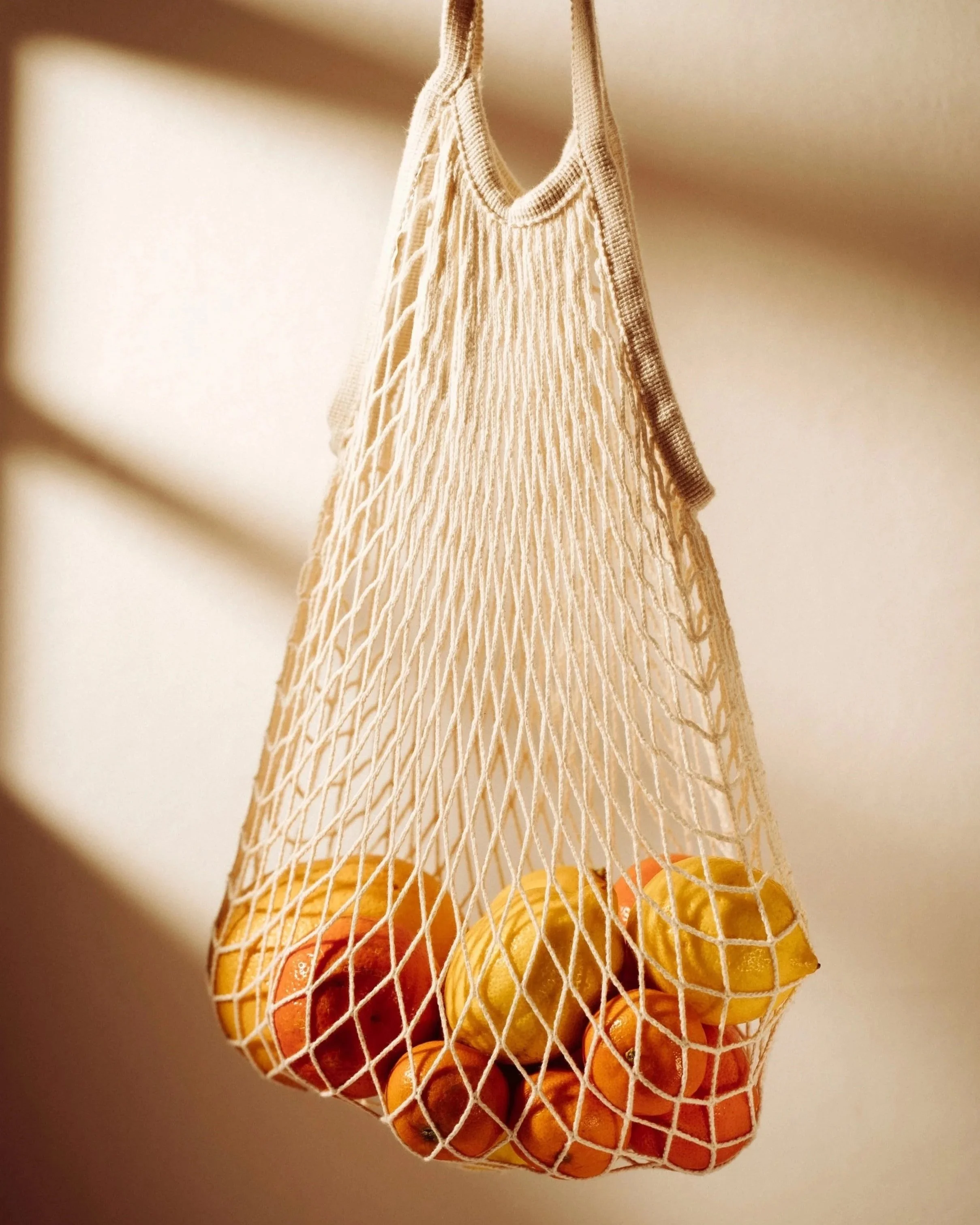 Mesh bag with oranges, lemons, and apples hanging against a beige background.