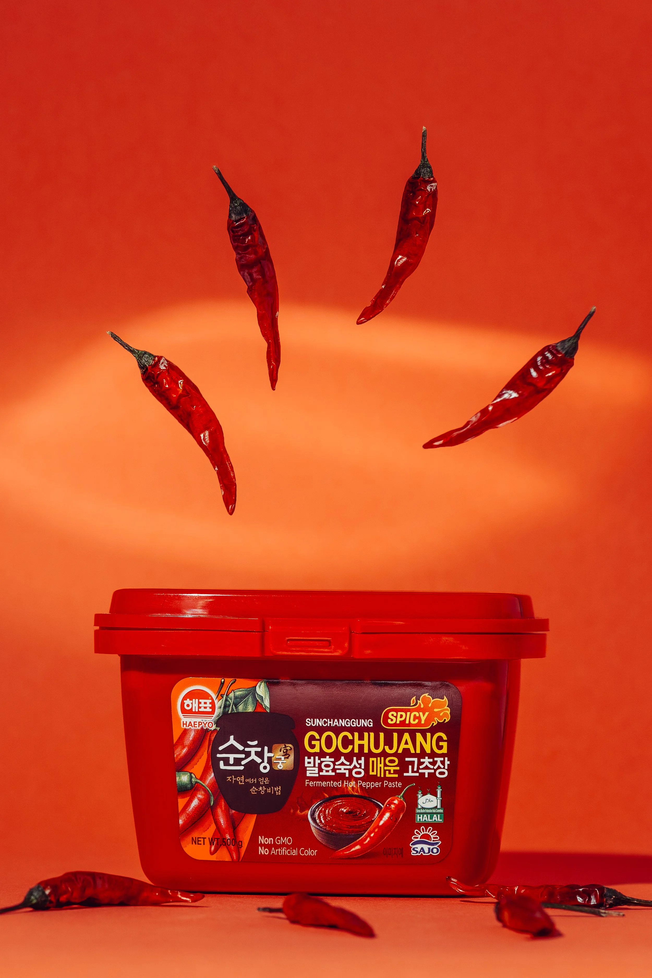 A red plastic container of fermented hot pepper paste with several red chili peppers floating above and around it against a red background.