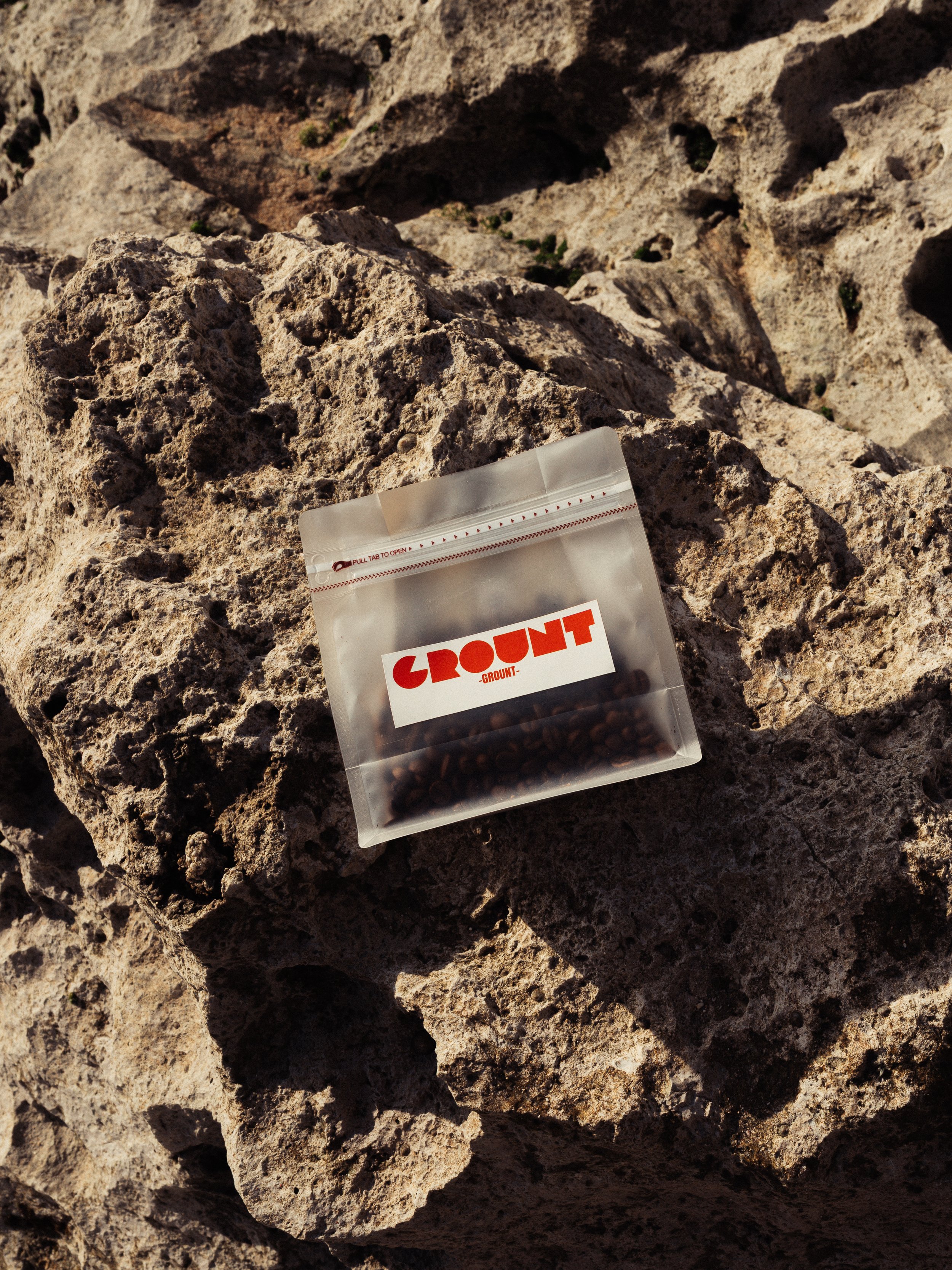 A partially opened transparent plastic bag with a red and white label reading 'CROUNT' is placed on a rough, textured rock surface with sun shadows.