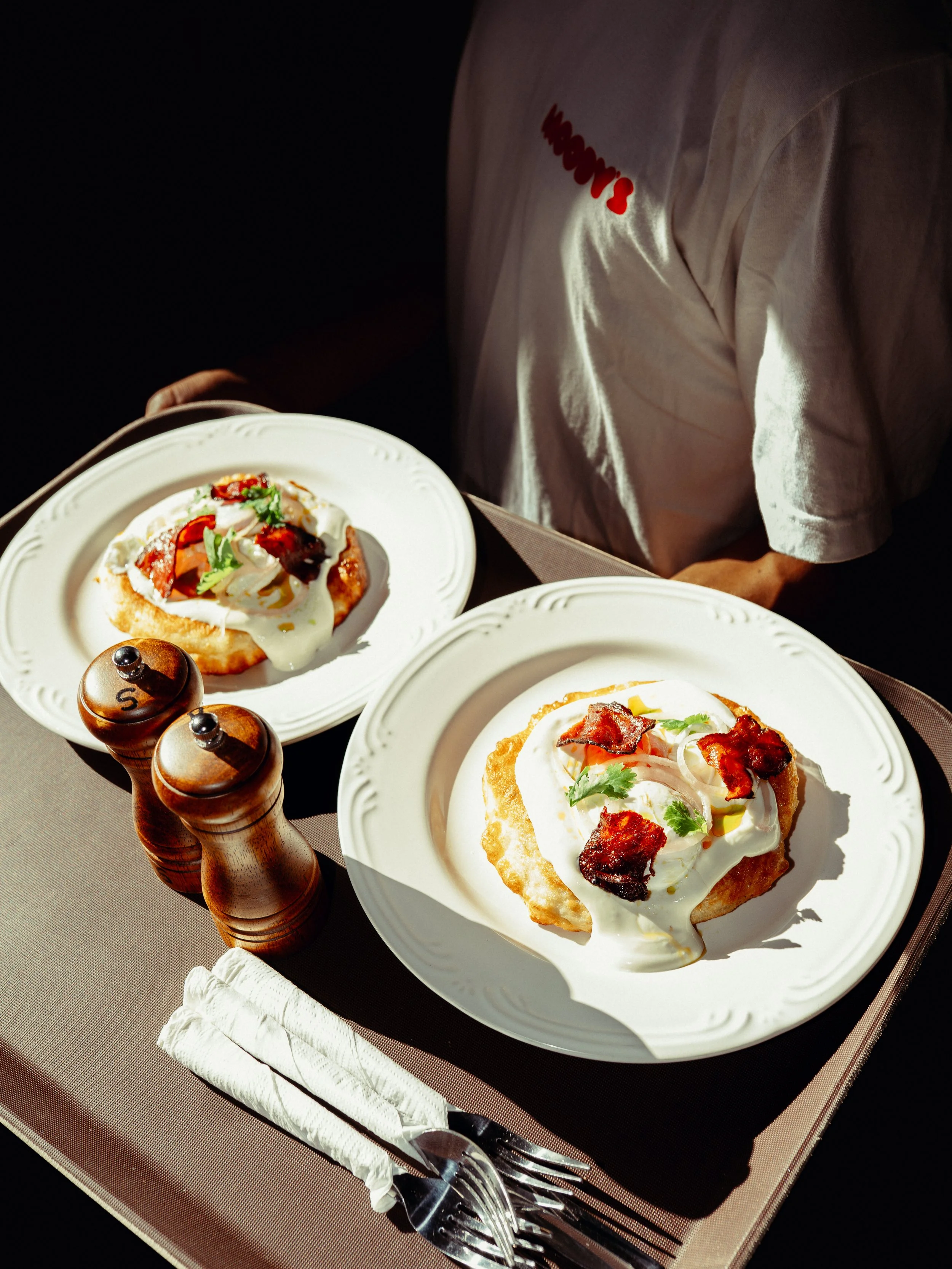 Plate of eggs Benedict with bacon, garnished with herbs, served with a side of potatoes, on a white plate, with salt and pepper shakers on a table.