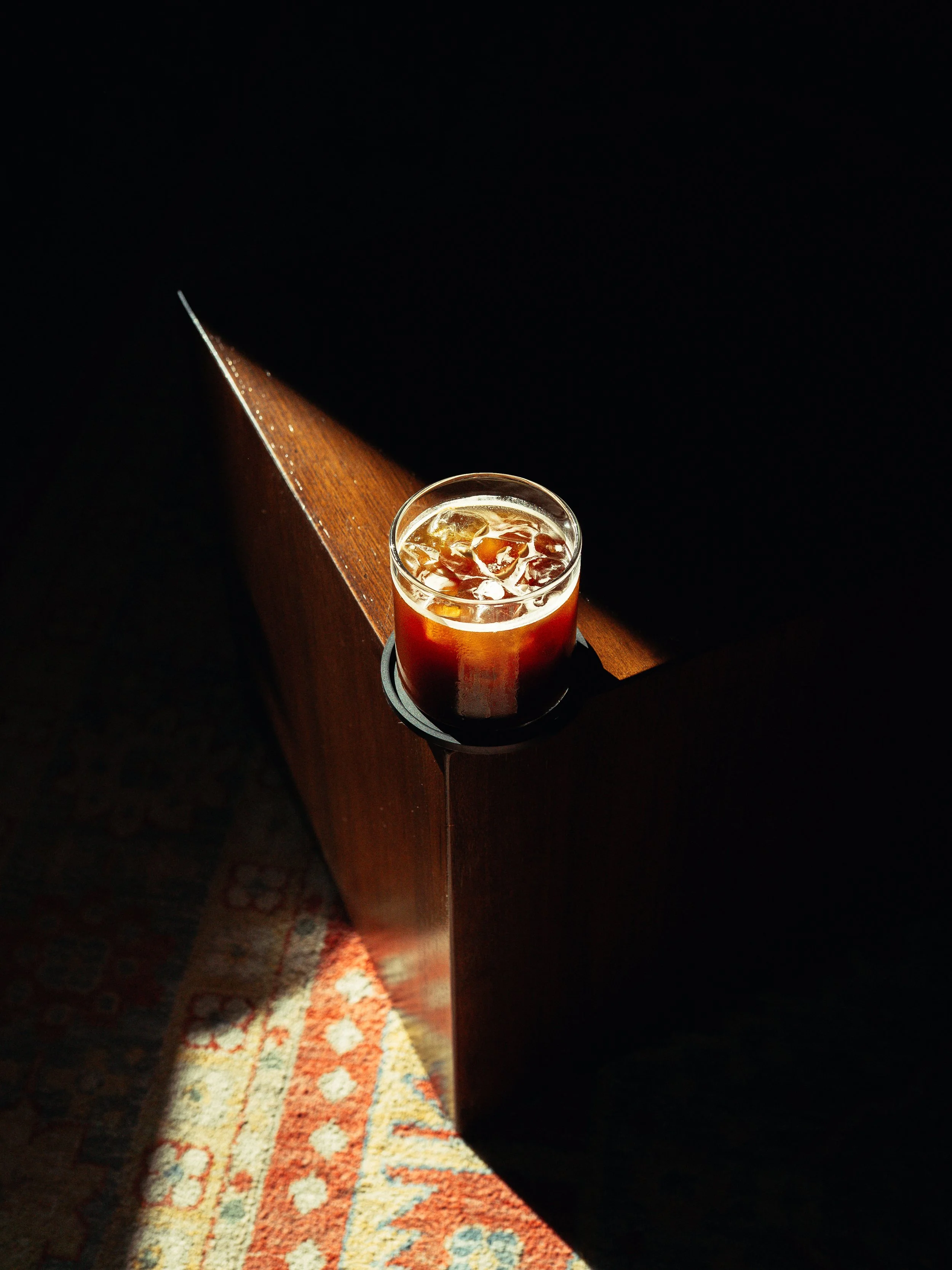 A glass of iced beverage on a wooden table with a dark background, and a patterned carpet visible on the floor.