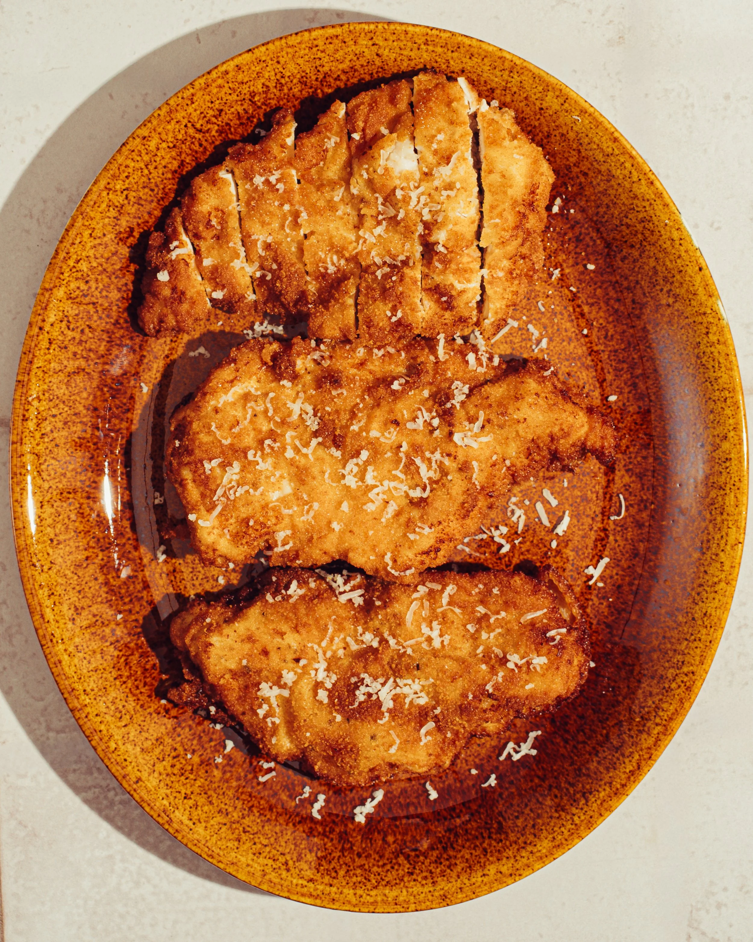 Fried chicken cutlets on a speckled orange plate, topped with grated cheese, on a white surface.