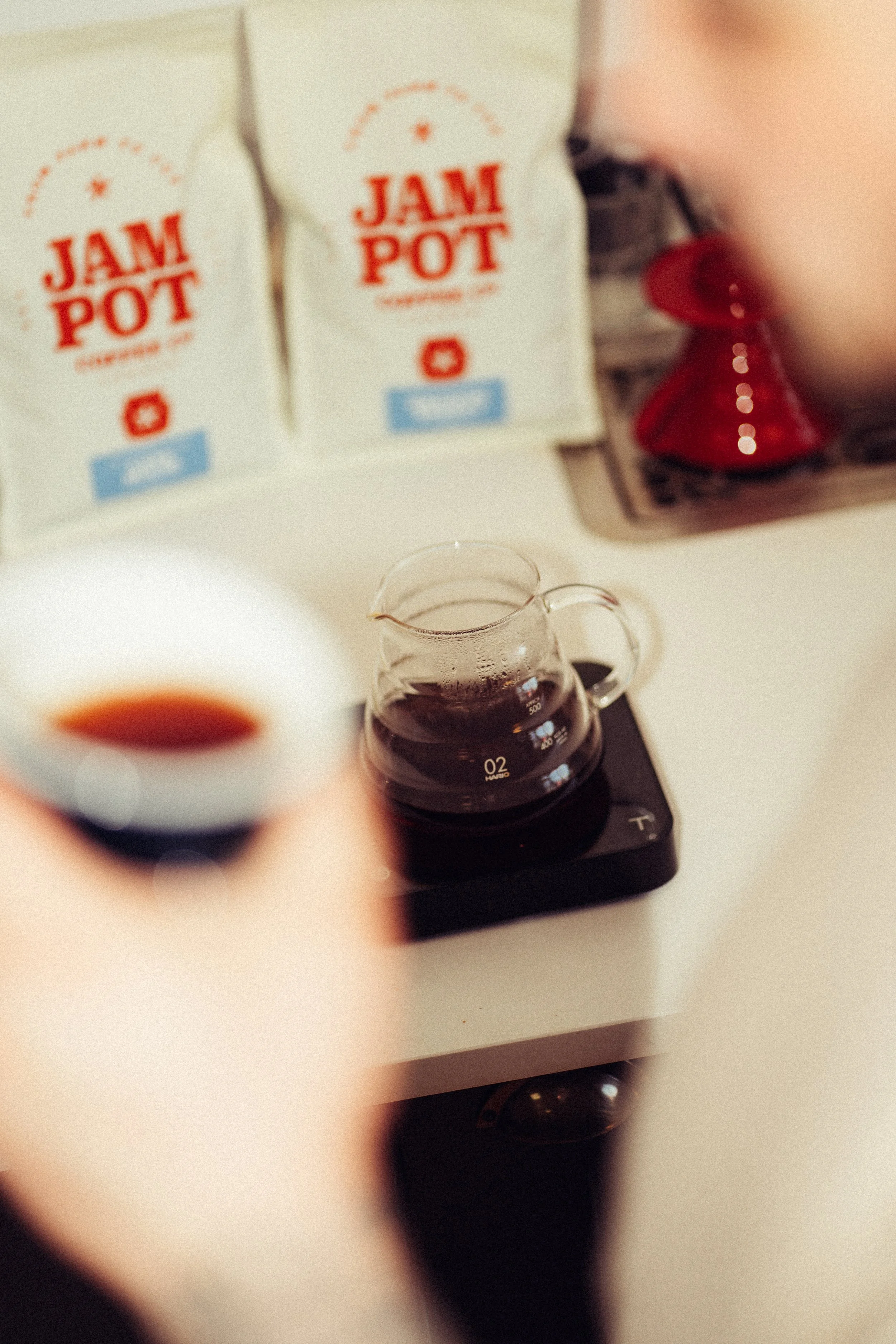 A steaming cup of coffee, a glass measuring cup filled with coffee on an electric stovetop, with a package of jam pot in the background.