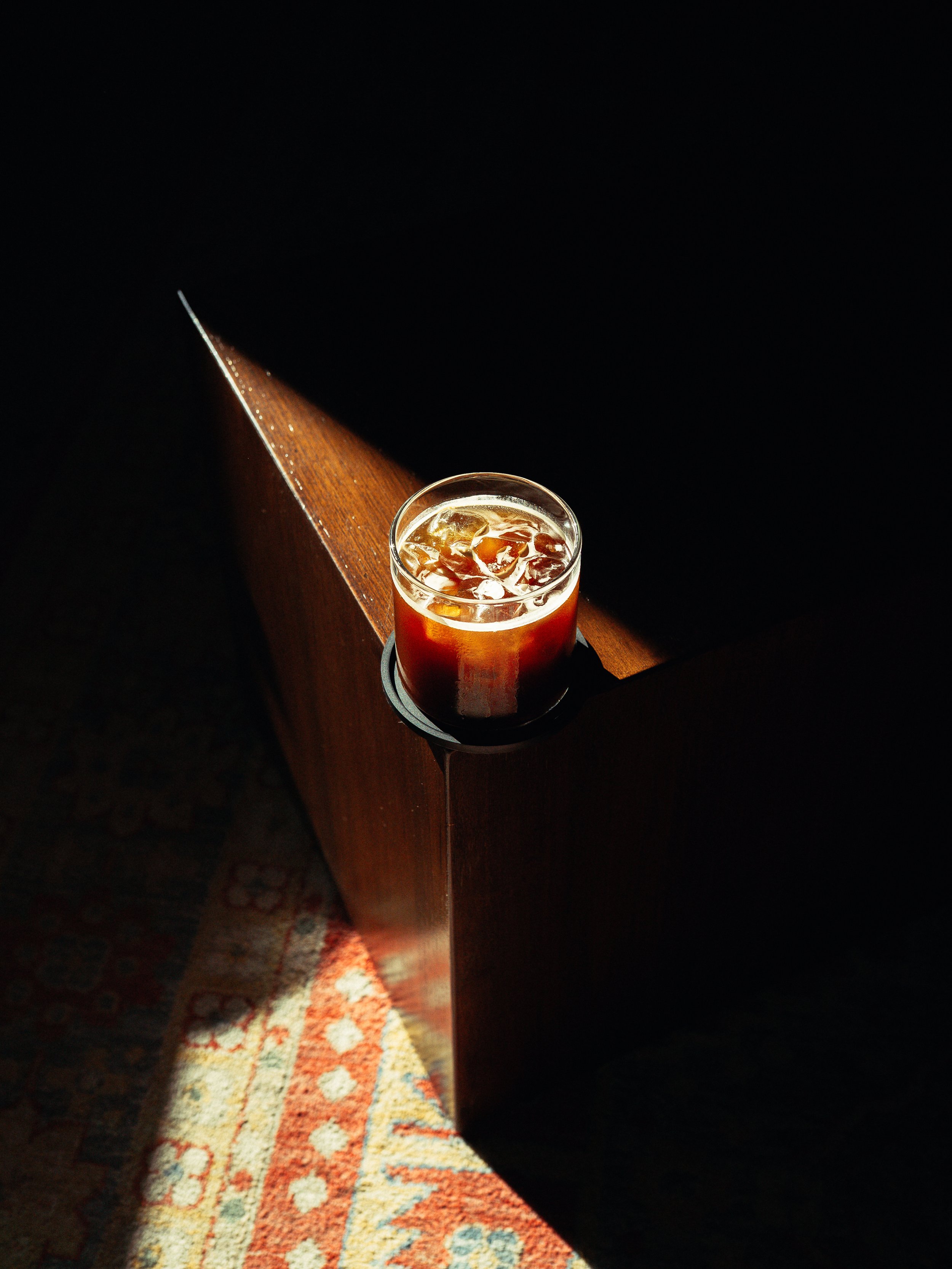 A glass of iced tea on a wooden surface with a dark background and a patterned rug visible in the lower left corner.