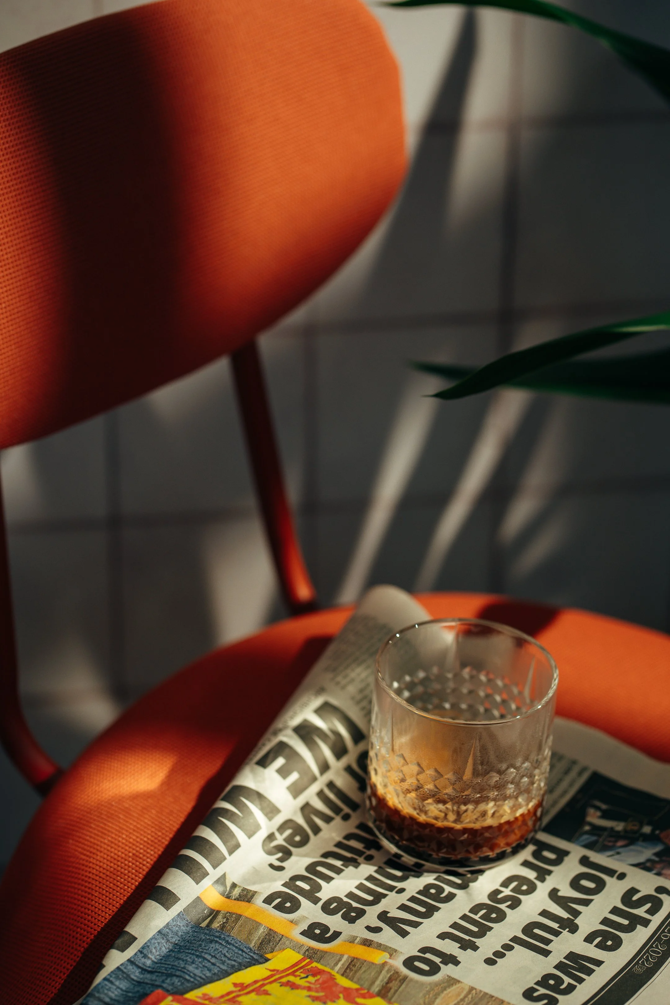 A glass of hot coffee, newspaper, an orange chair, and green indoor plant leaves in a cozy setting with sunlight.