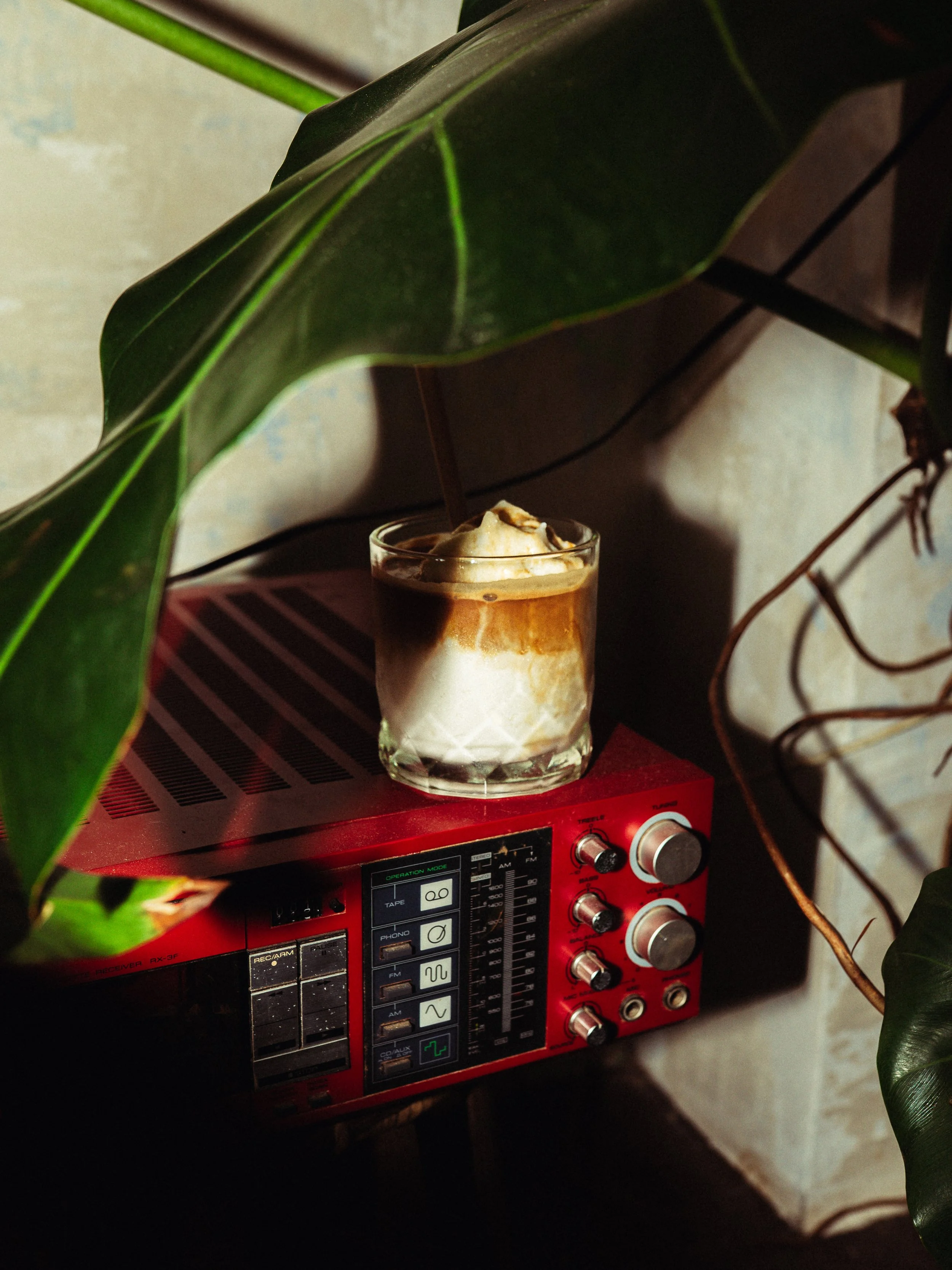 A glass of iced coffee placed on top of a red vintage radio and a small table. The coffee has whipped cream and caramel syrup on top. Large green leaves partially cover the scene.