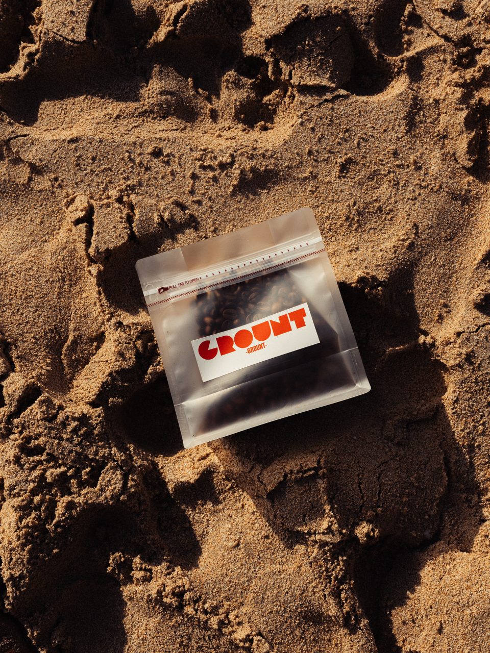 A clear plastic bag of coffee beans with a white and orange 'CROUNT' label, lying on sandy ground.