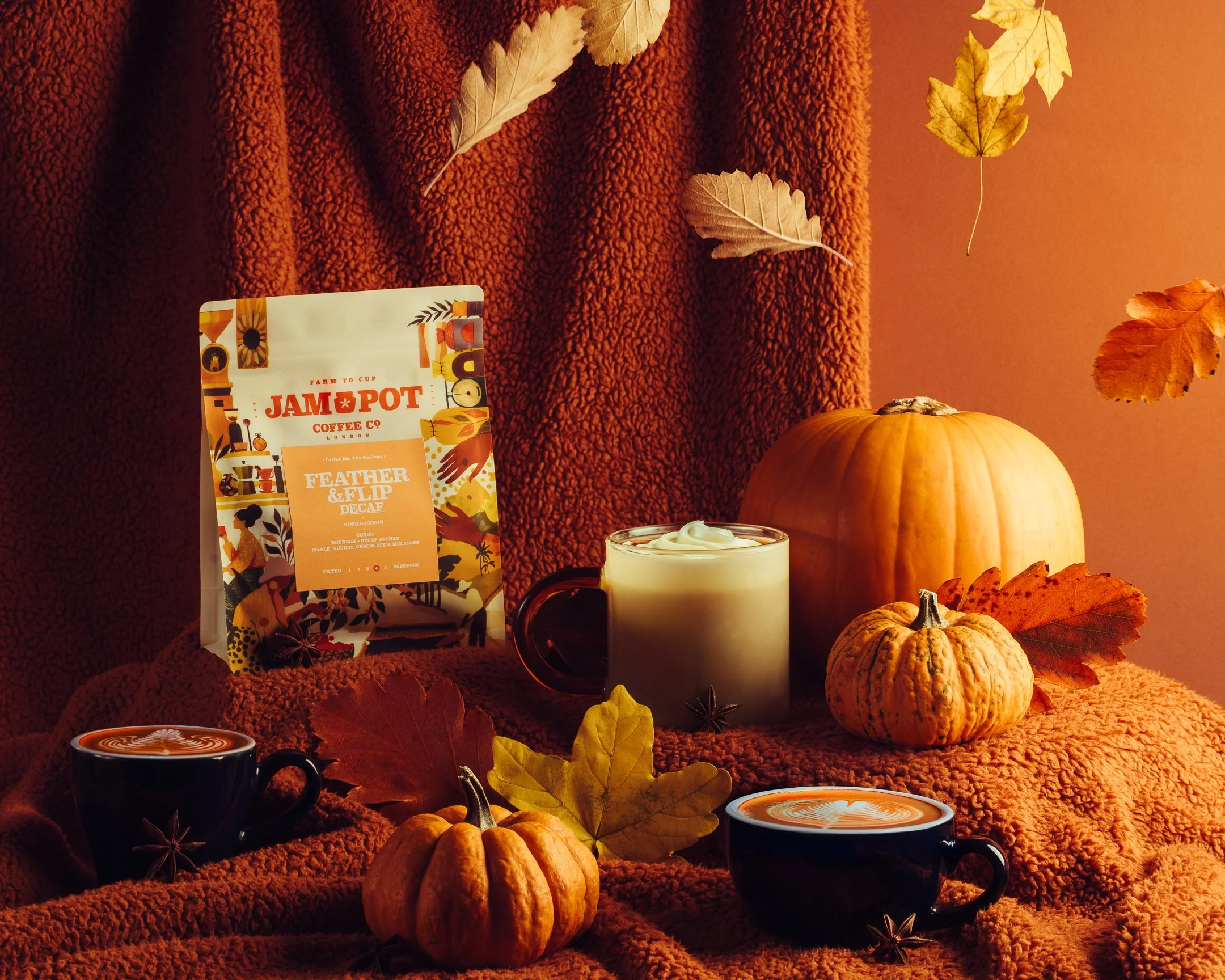 Autumn scene with pumpkins, fall leaves, coffee cups with latte art, a bag of coffee, and a jar of whipped cream on a cozy, textured orange blanket.