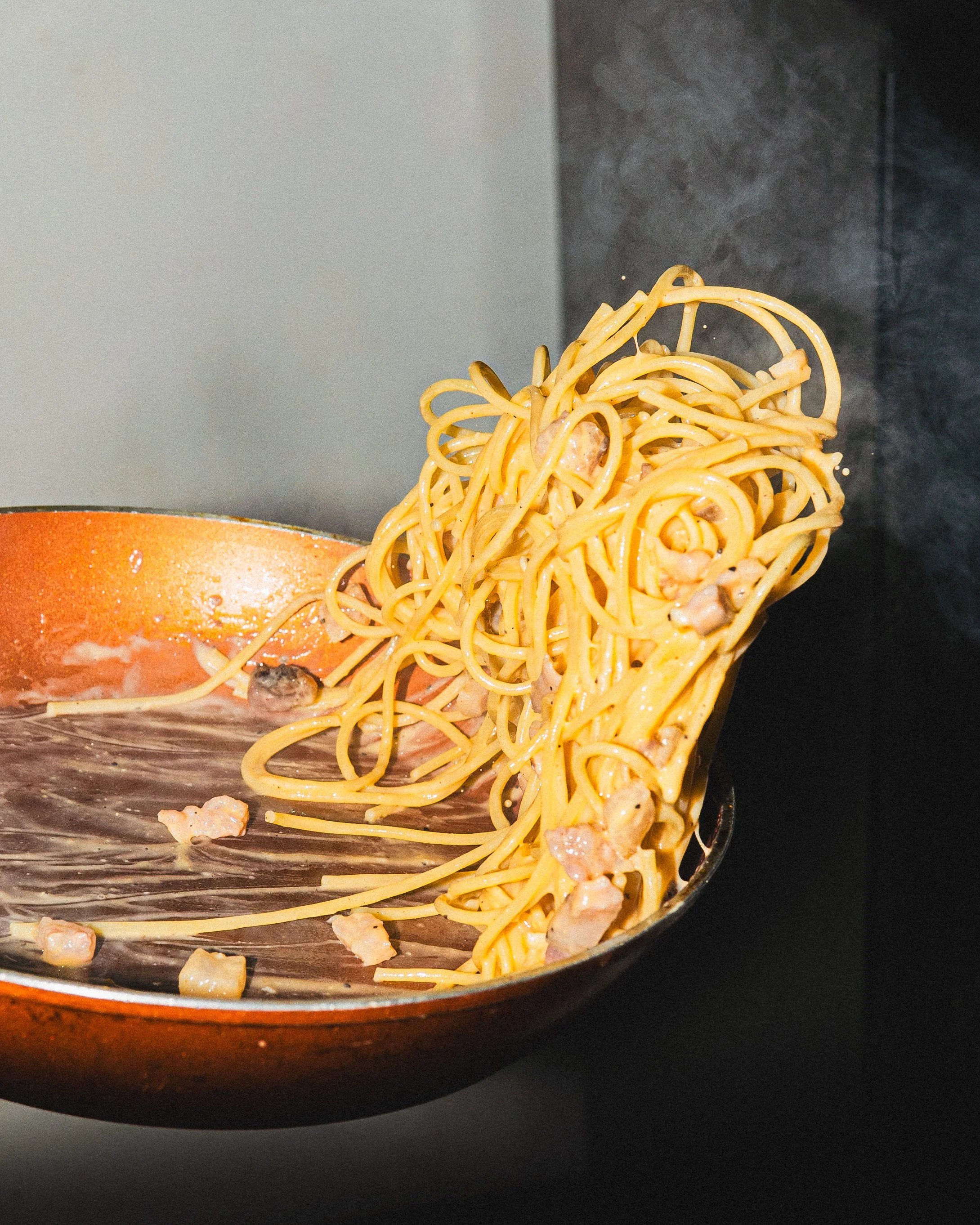 Close-up of spaghetti being lifted with a fork from a skillet, with some pieces of bacon visible, on a black background.
