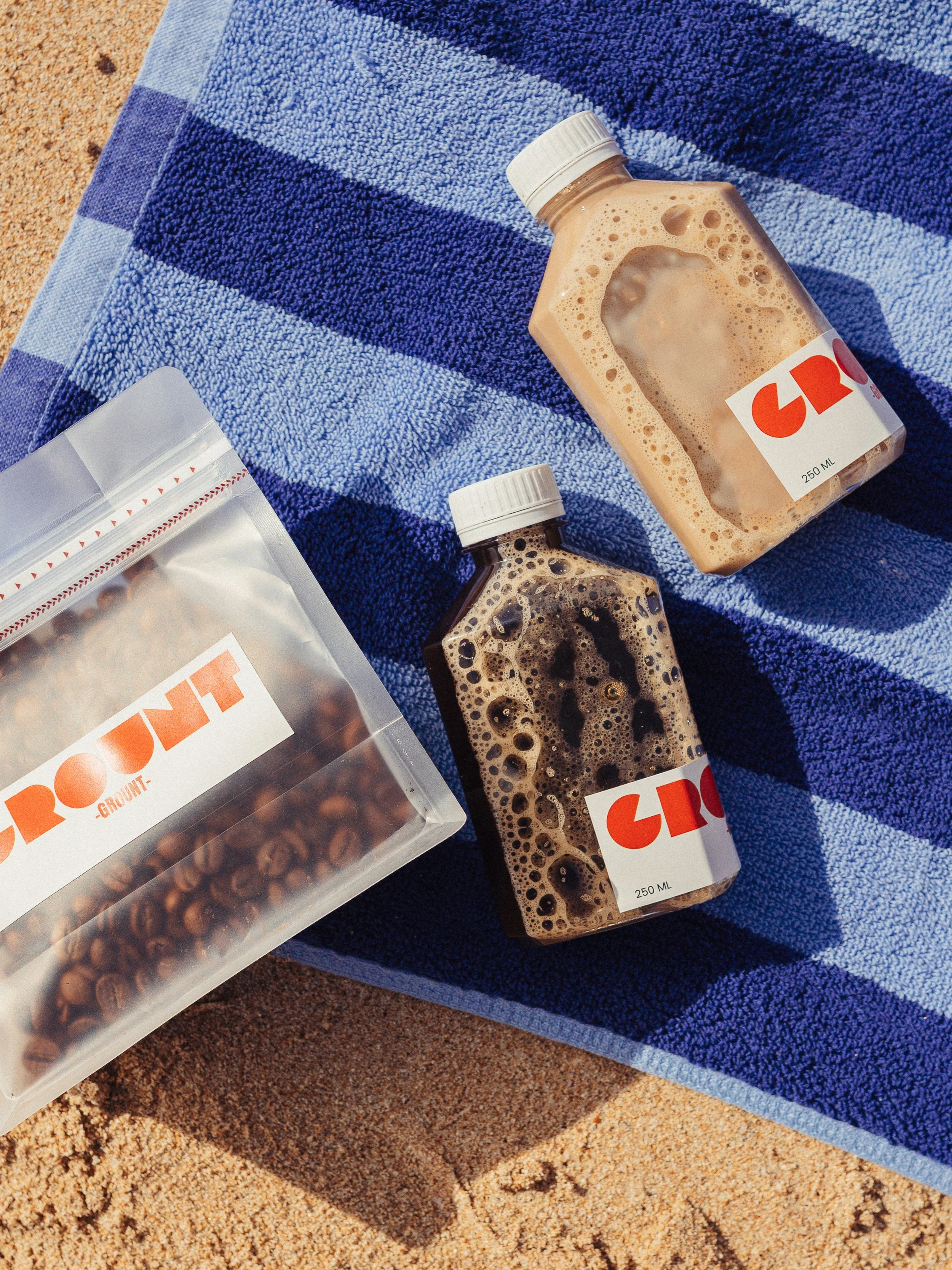 Two bottles of iced coffee or chocolate drinks on a blue towel on sandy ground, next to a clear plastic bag of coffee beans.