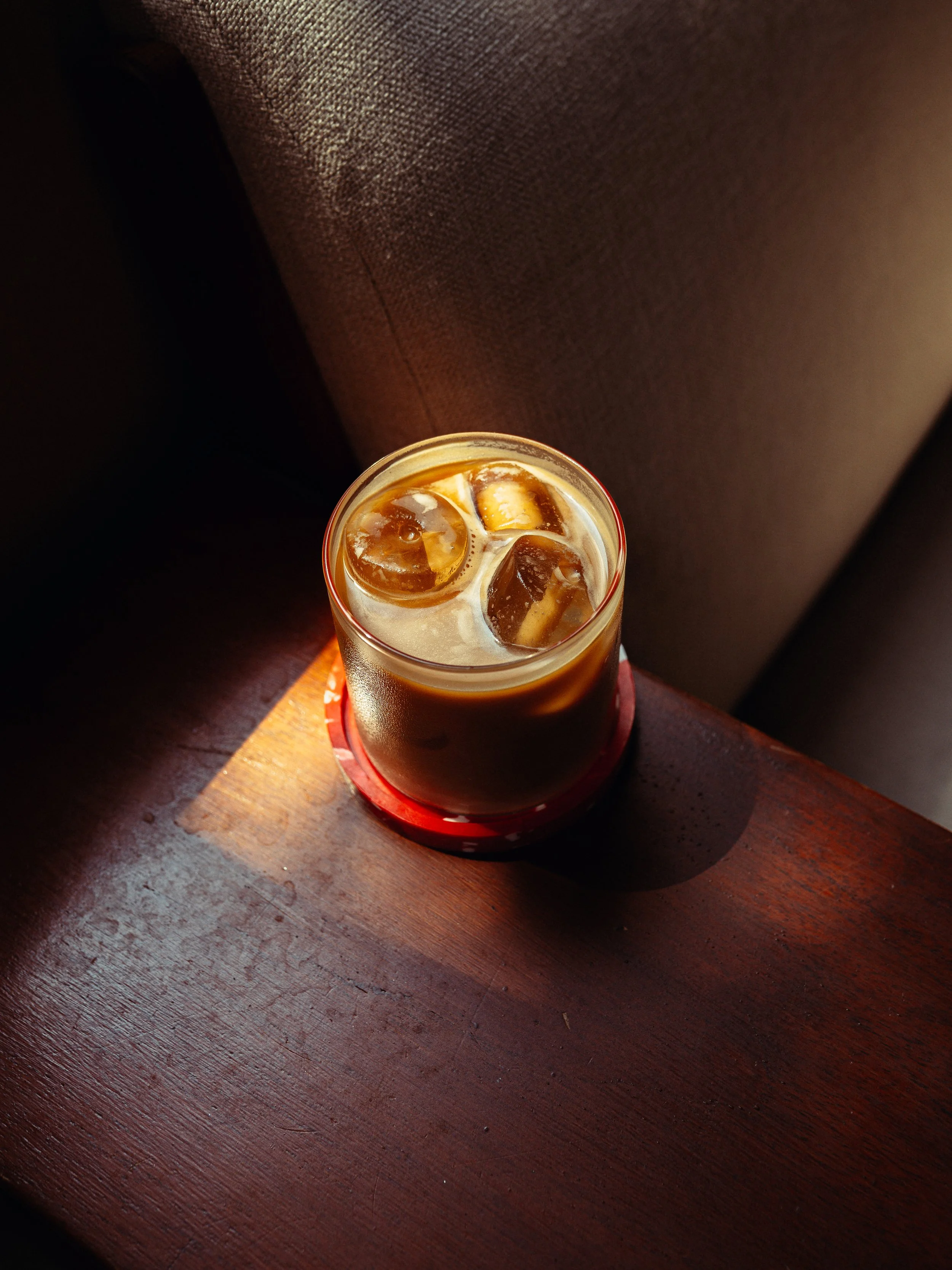 A glass of iced coffee with ice cubes sitting on a wooden table near a brown upholstered chair.