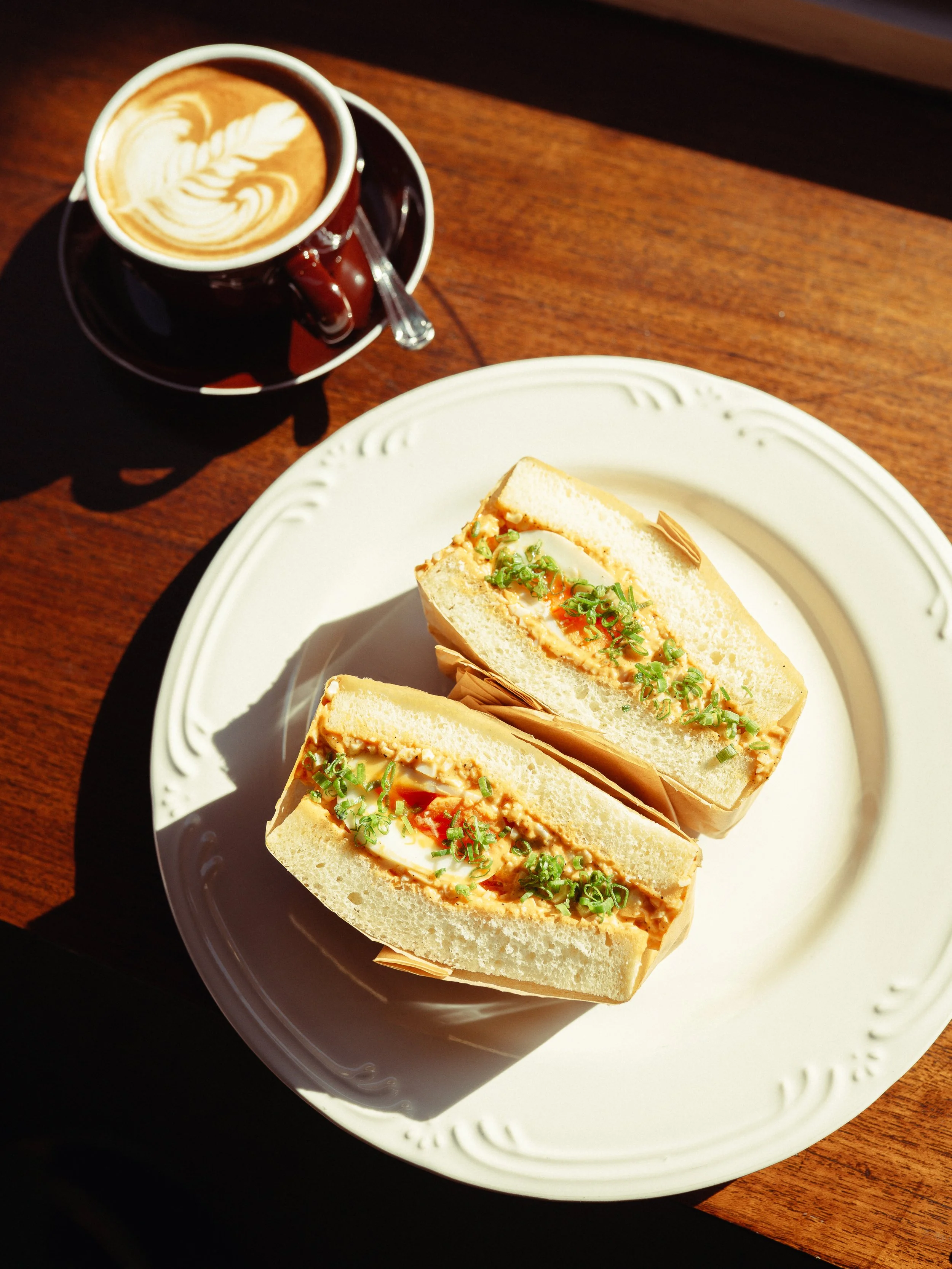 Close-up of a white plate with two club sandwiches filled with egg salad and garnished with chopped green onions, placed on a wooden table, with a cup of coffee topped with latte art nearby.