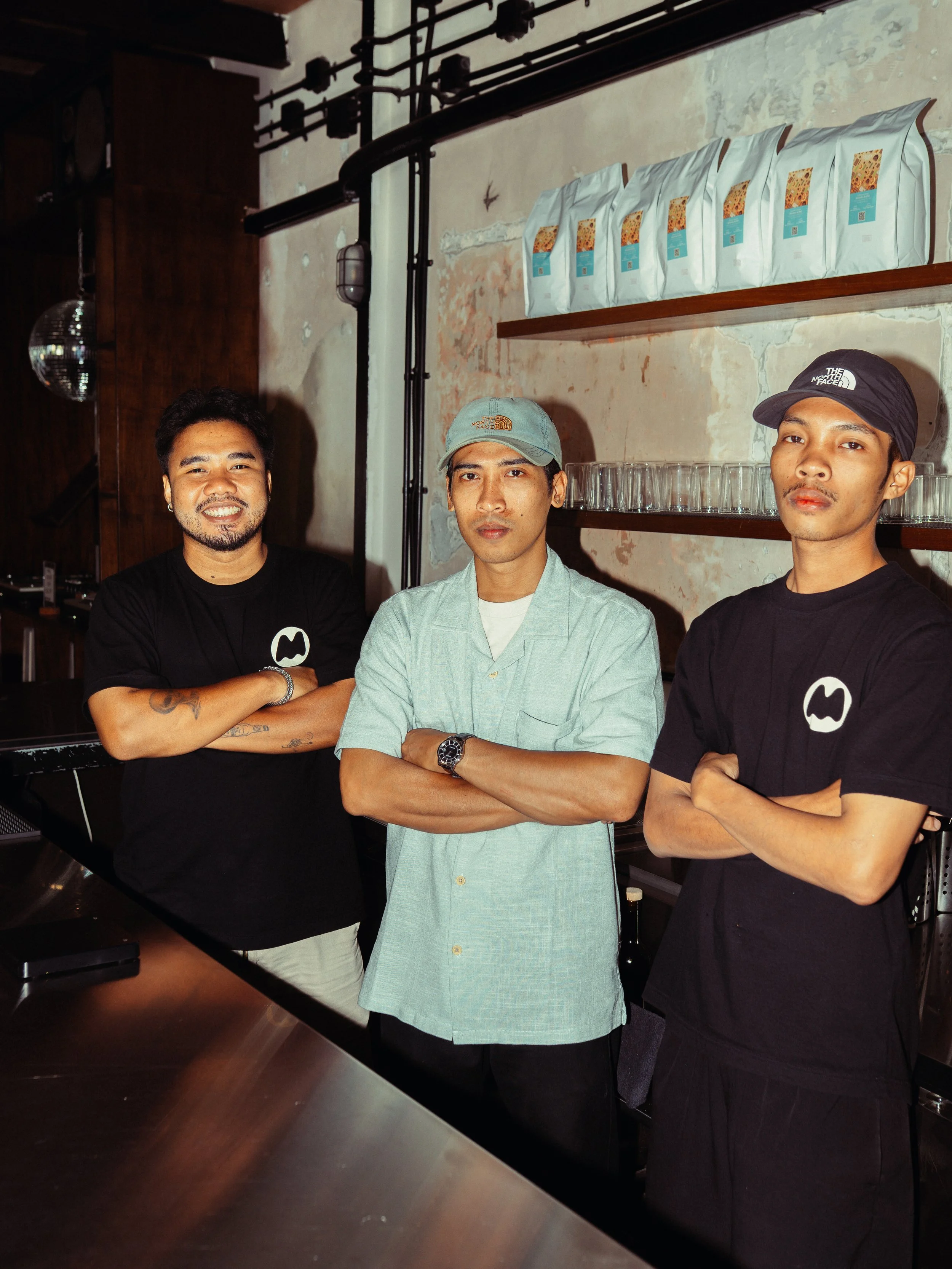 Three men standing with crossed arms in a dimly lit room with a rustic industrial style. One man is wearing a black T-shirt, another is wearing a light green shirt and cap, and the third is wearing a black T-shirt and cap. There are shelves with coff
