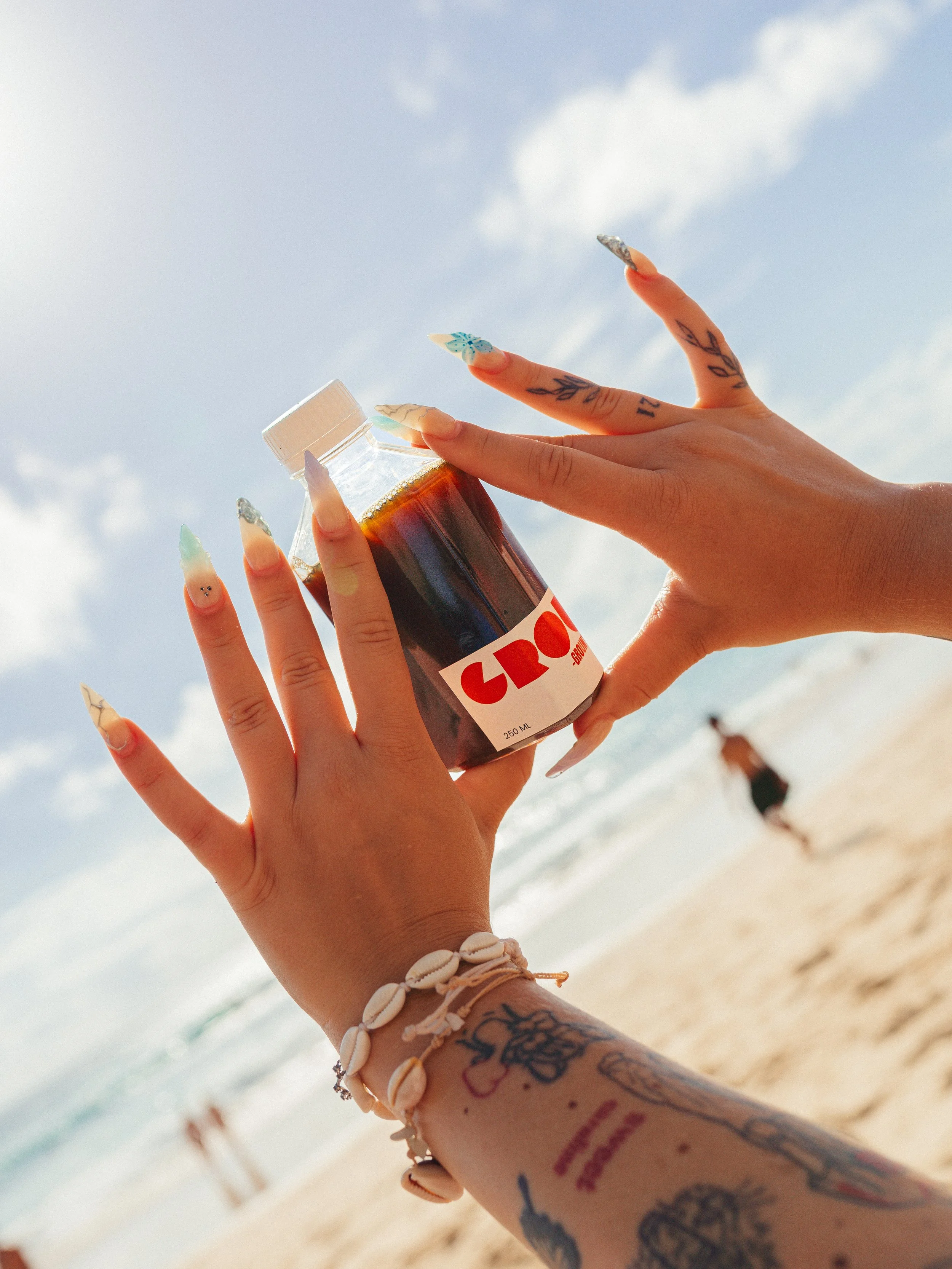 Person with tattooed arm holding a bottle of Cold brew at the beach with a cloudy sky and blurred person in the background.