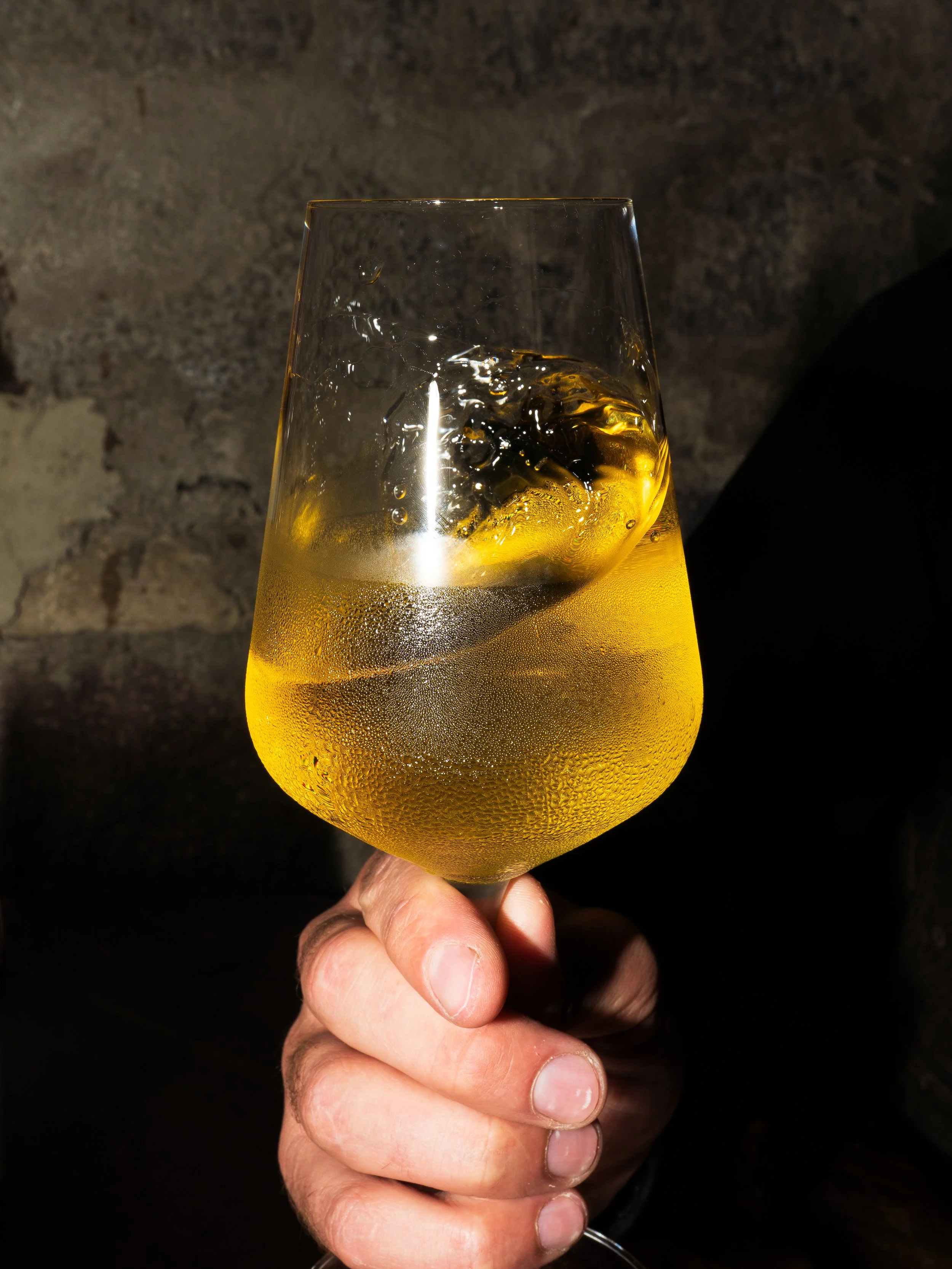 Close-up of a hand holding a glass of white wine with condensation on the outside, against a dark, textured background.