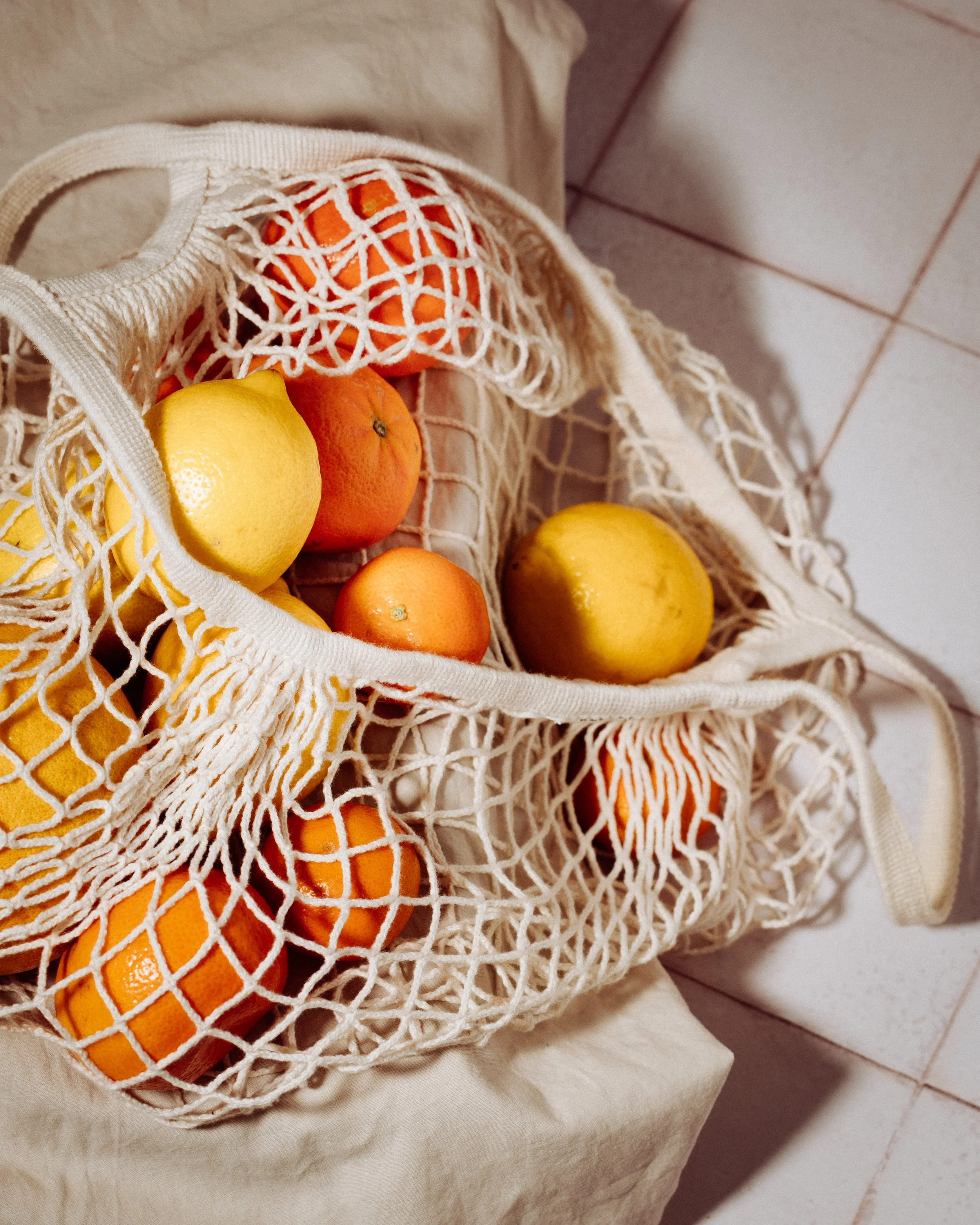 A beige reusable mesh bag filled with oranges and lemons, resting on a white fabric surface next to a tiled floor.
