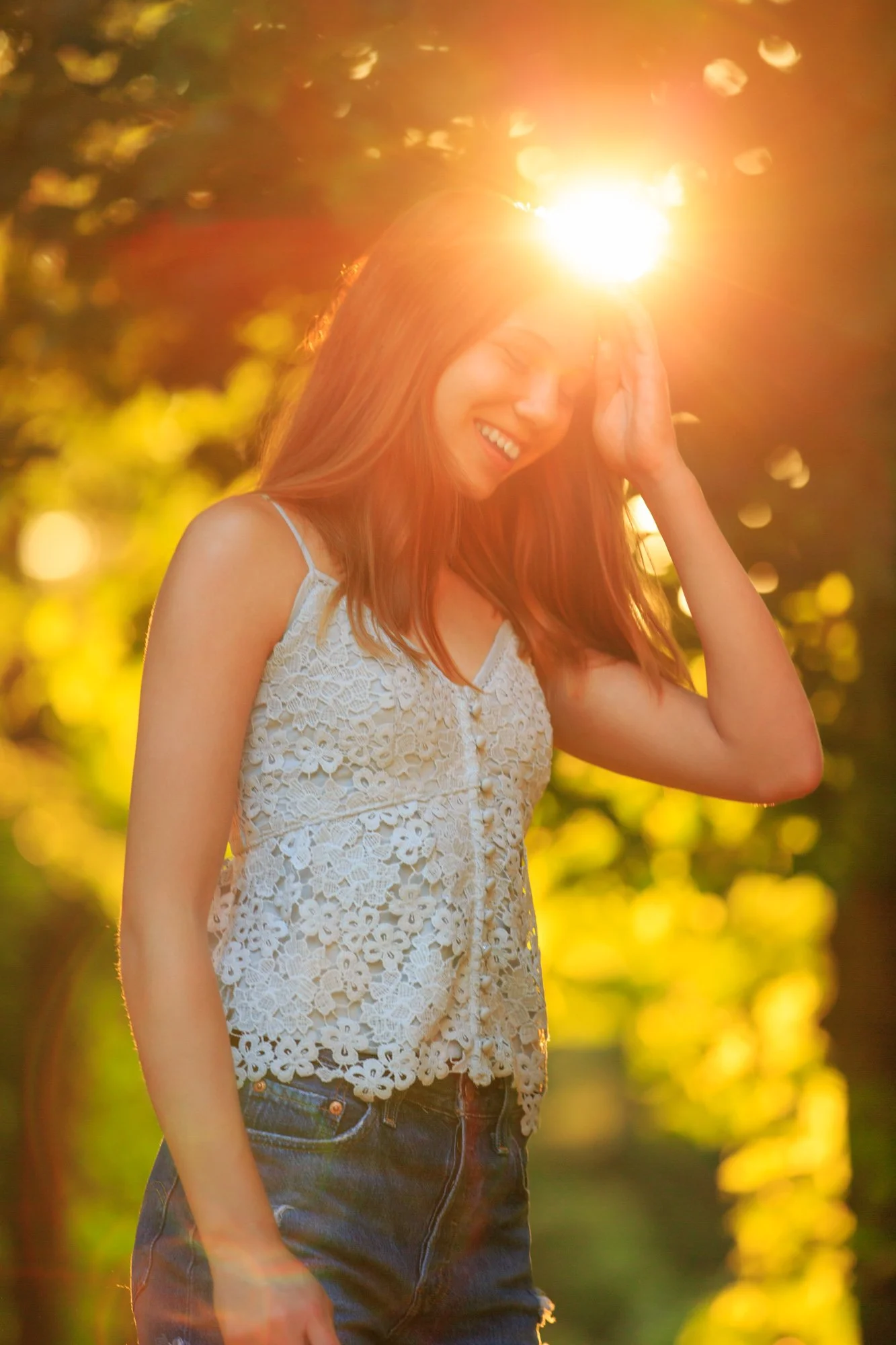 Smiling woman in lace top and denim shorts with sunlight filtering through trees behind her.