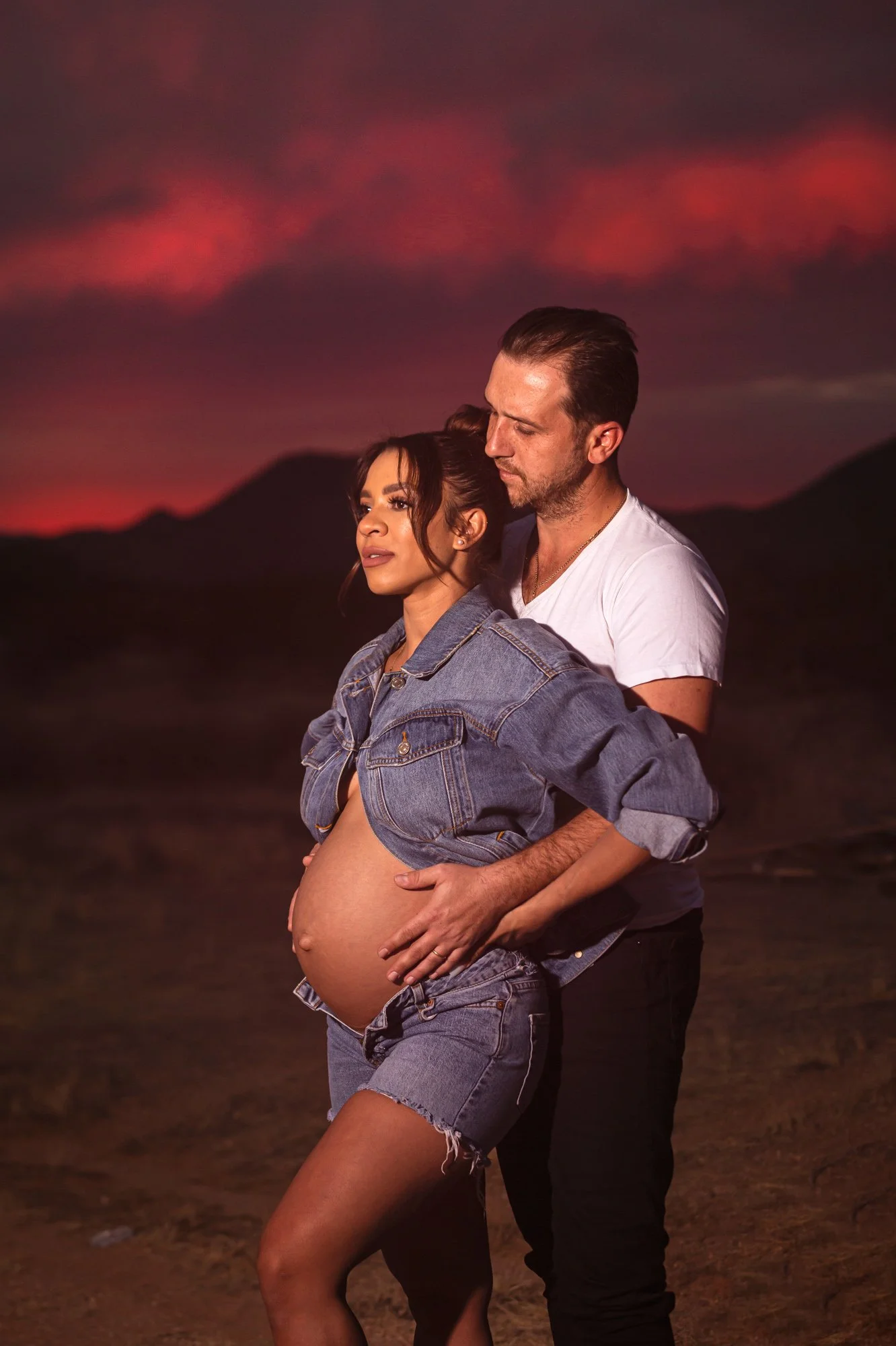 Couple embracing during sunset in desert landscape; woman showing pregnancy.