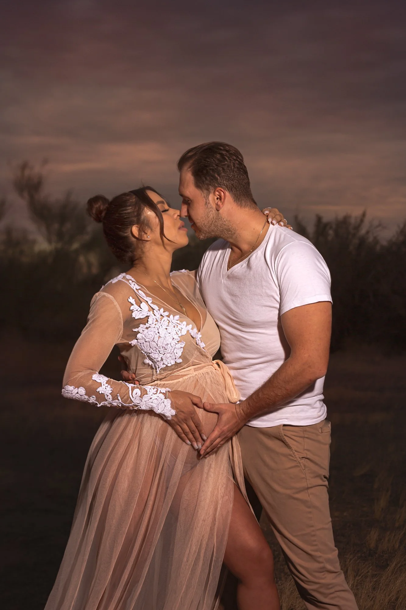 A couple posing in an embrace outdoors at sunset, with the man in a white shirt and the woman in a beige, sheer dress with lace details. They form a heart shape with their hands over her belly, suggesting a maternity photo shoot.