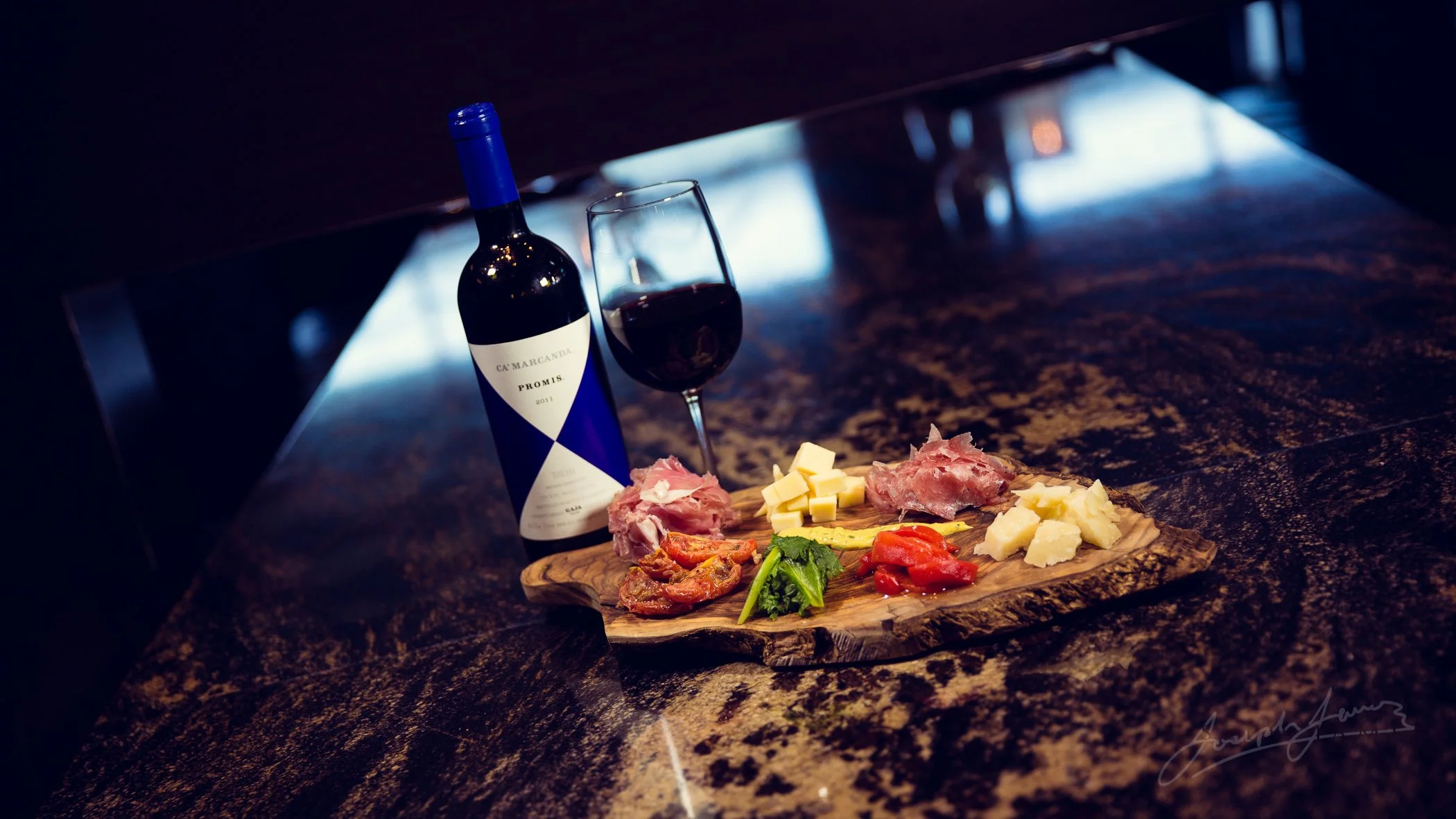 Bottle of red wine, glass of wine, charcuterie board with cured meats, cheeses, and vegetables on wooden table.
