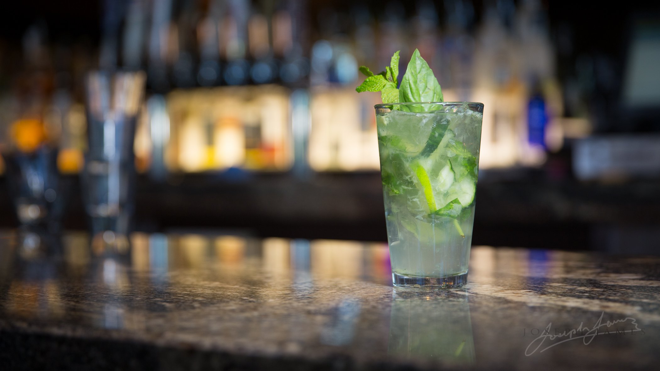 Glass of mojito cocktail on a bar counter, garnished with mint leaves and lime slices, with blurred bottles in the background.