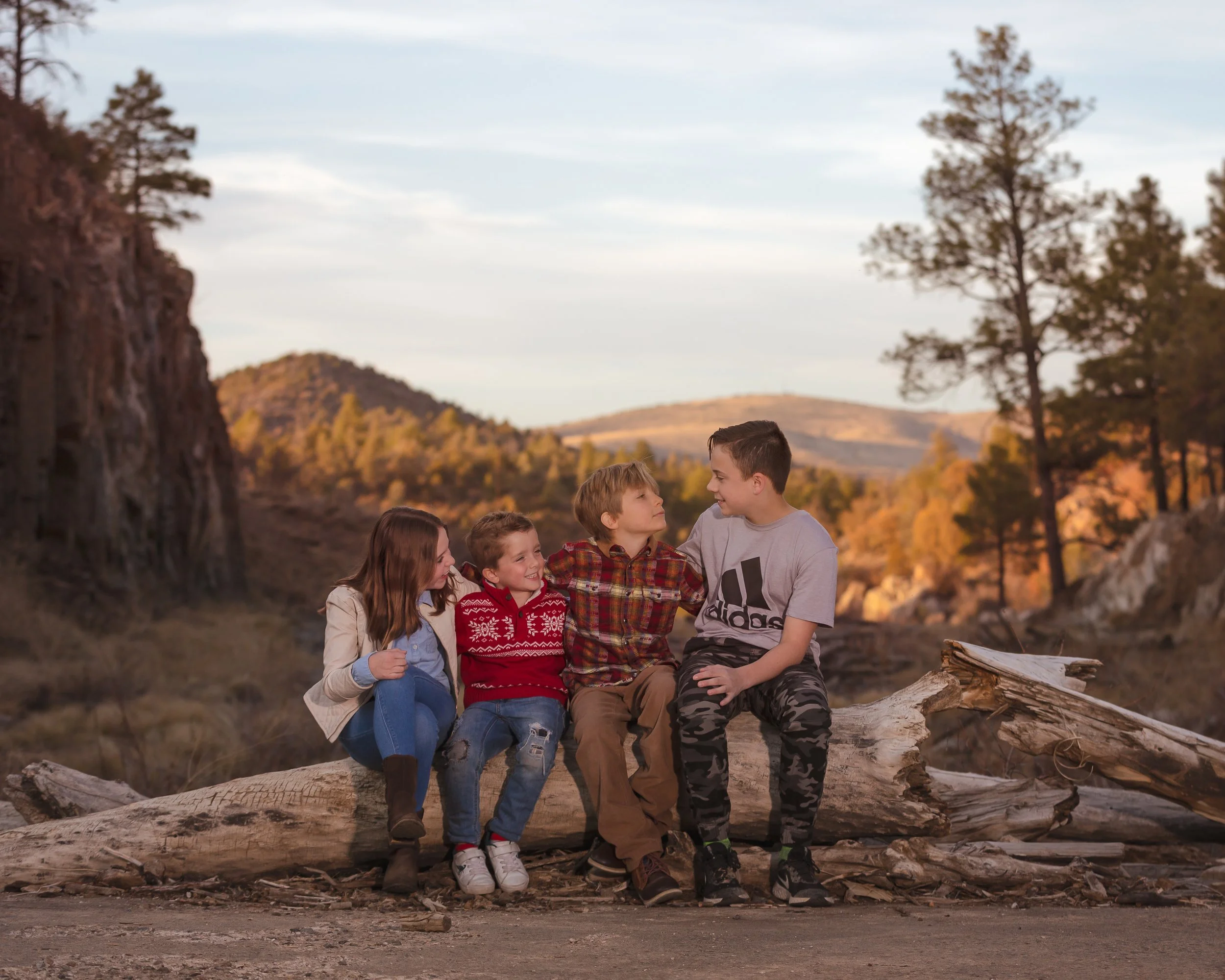 A group of four children sitting on a large log in a natural outdoor setting, with trees and hills in the background.
