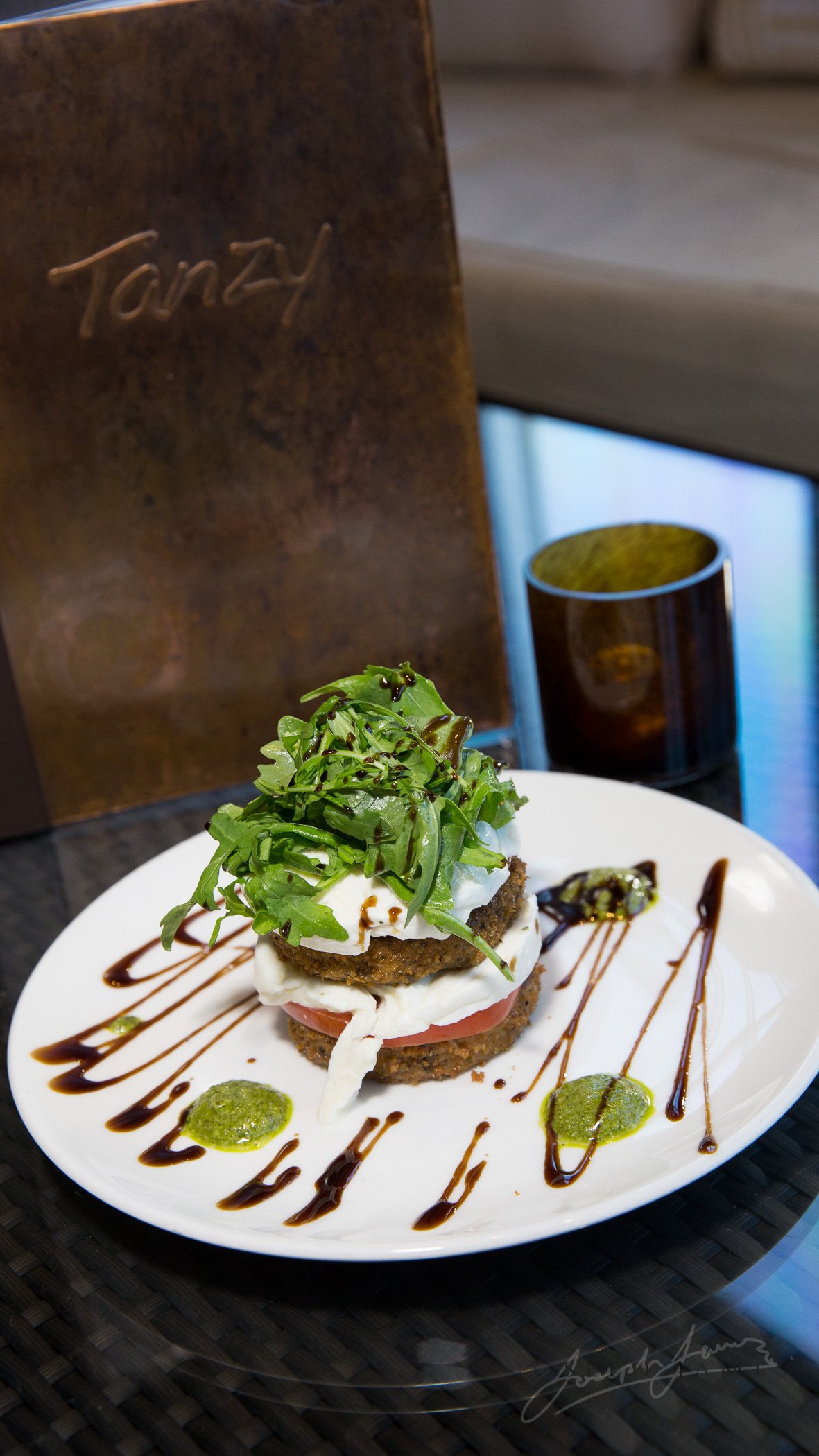 A gourmet plate featuring a layered dish with greens, cheese, tomato, and a patty. Drizzled with balsamic glaze and green sauce, set on a white plate next to a brown menu and glass.