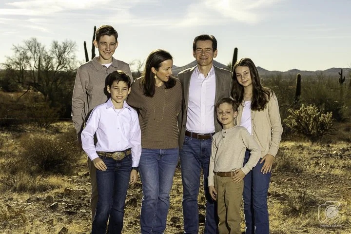 A group of six people posing outdoors, consisting of two adults and four children, standing together on a dry, desert landscape with cacti in the background.