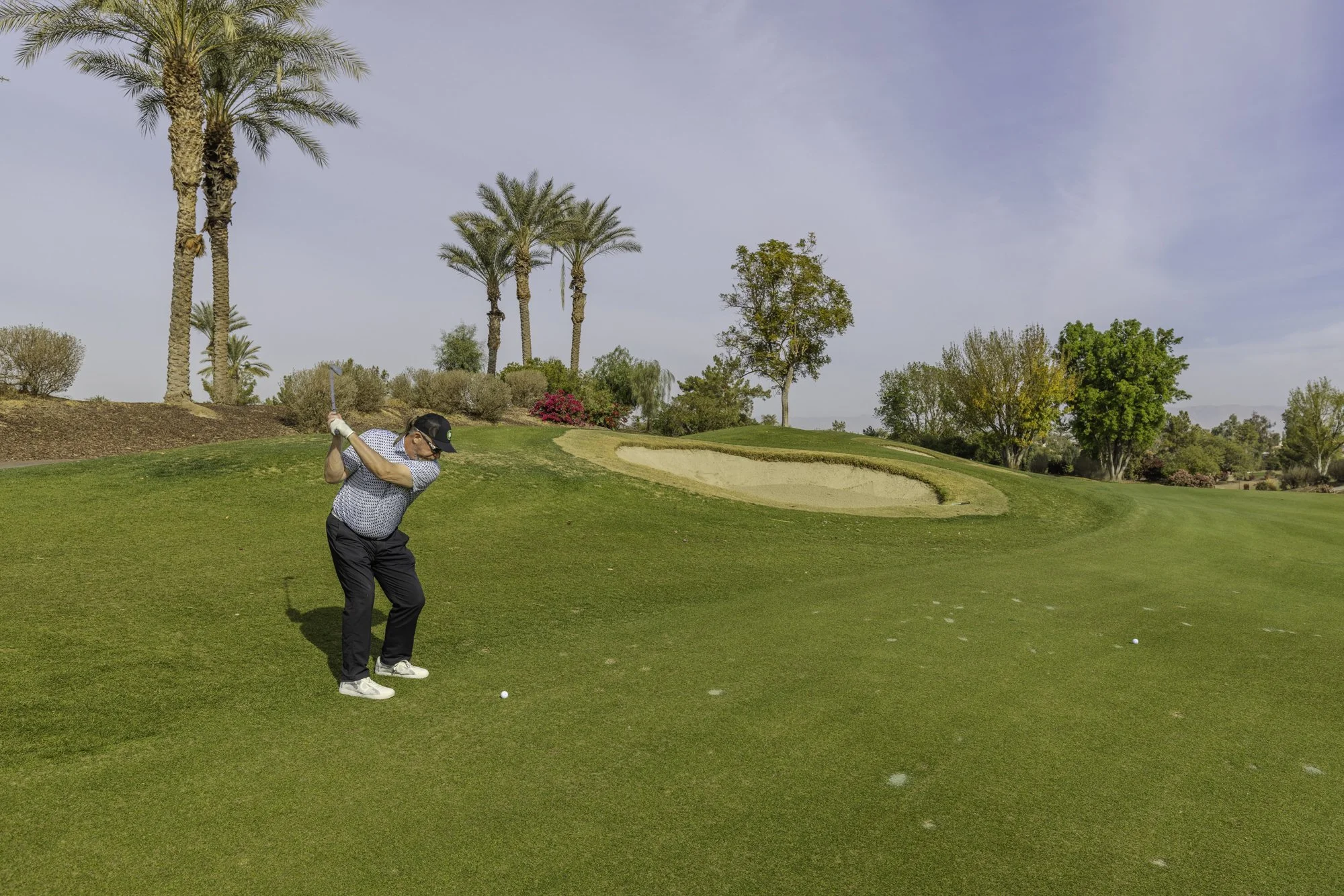 Golfer swinging club on lush green golf course, sand bunker and palm trees in background.