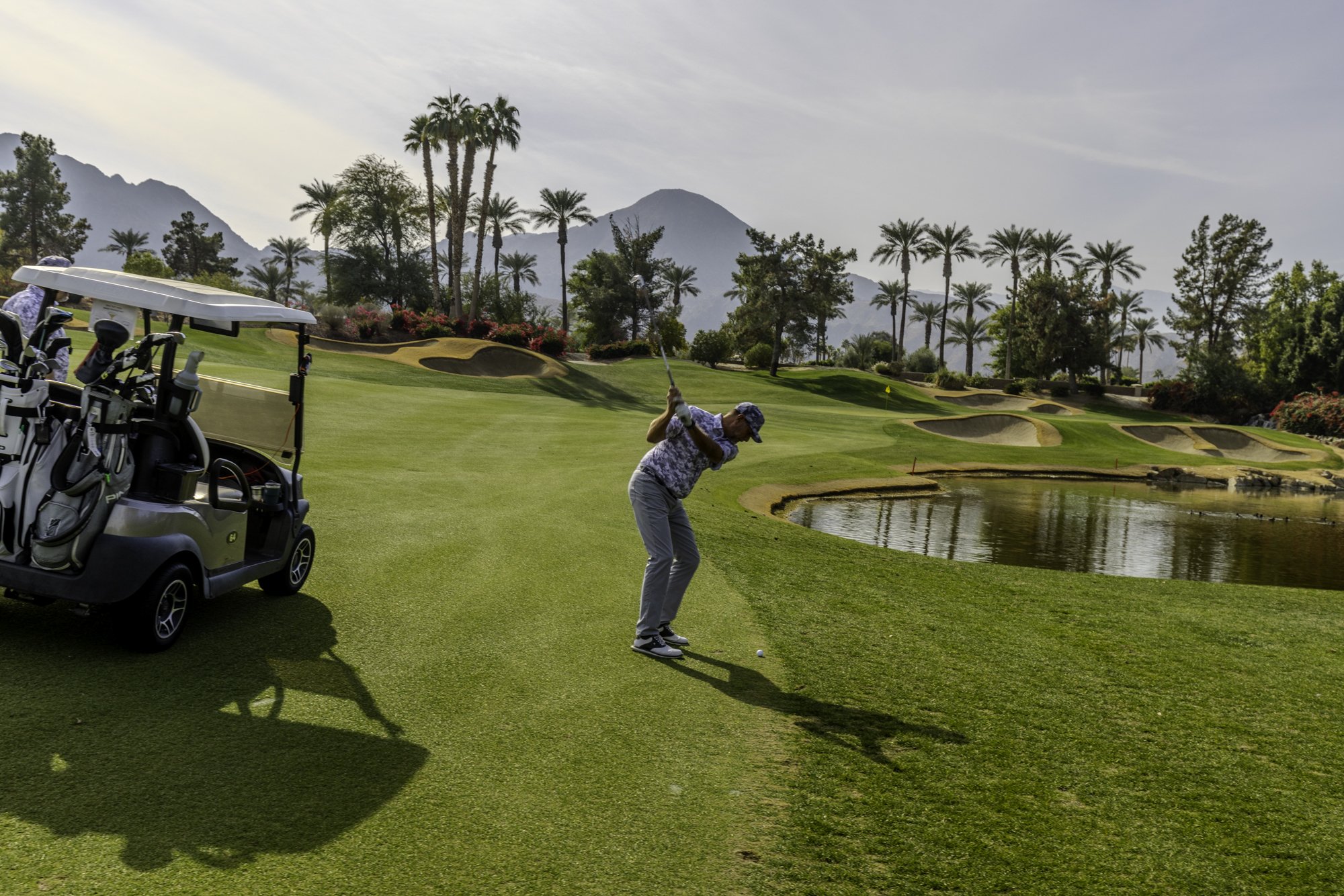 Golfer swinging club on green golf course near water, palm trees and mountains in the background.
