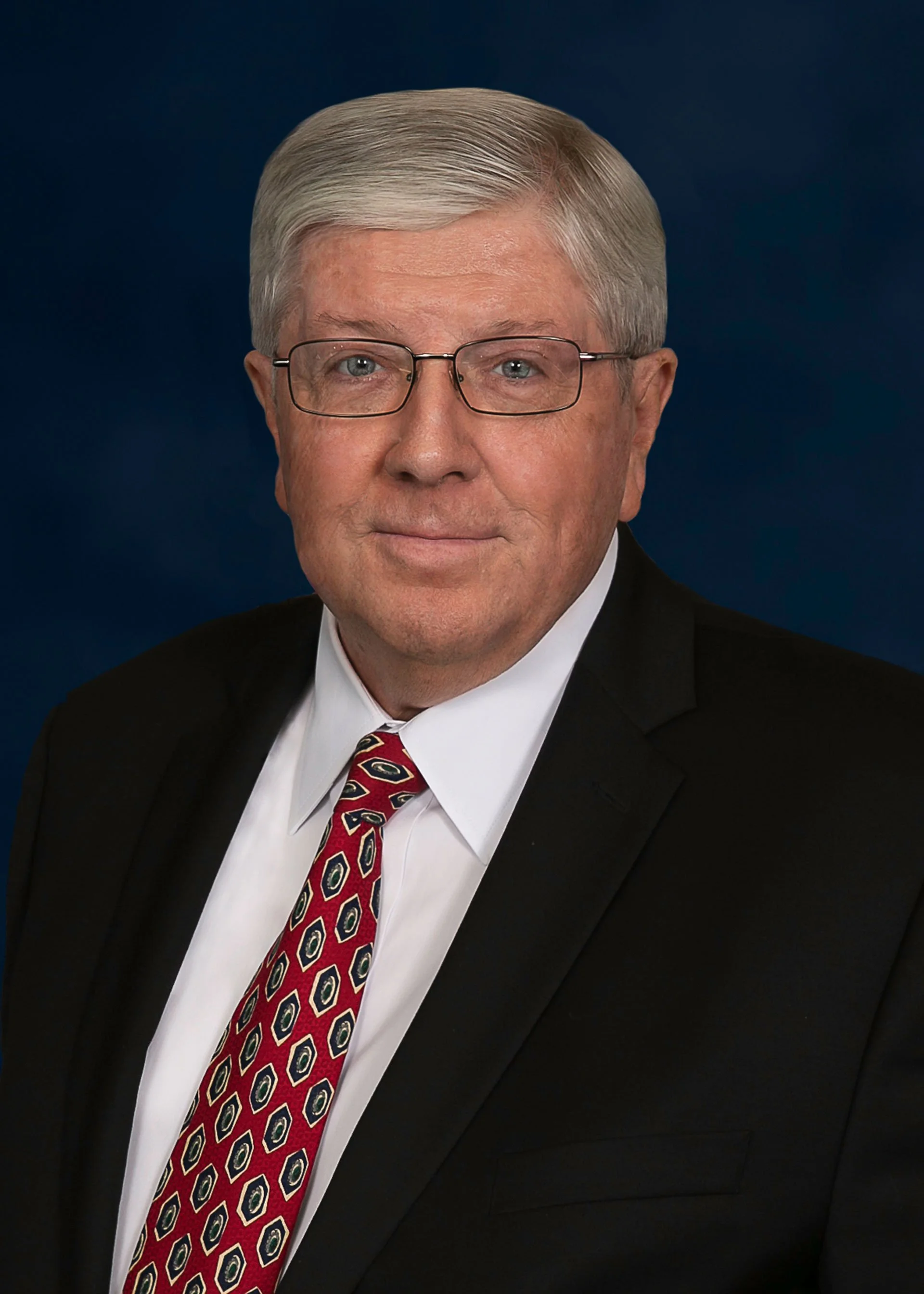 Man in glasses wearing a suit and patterned tie with a dark background.