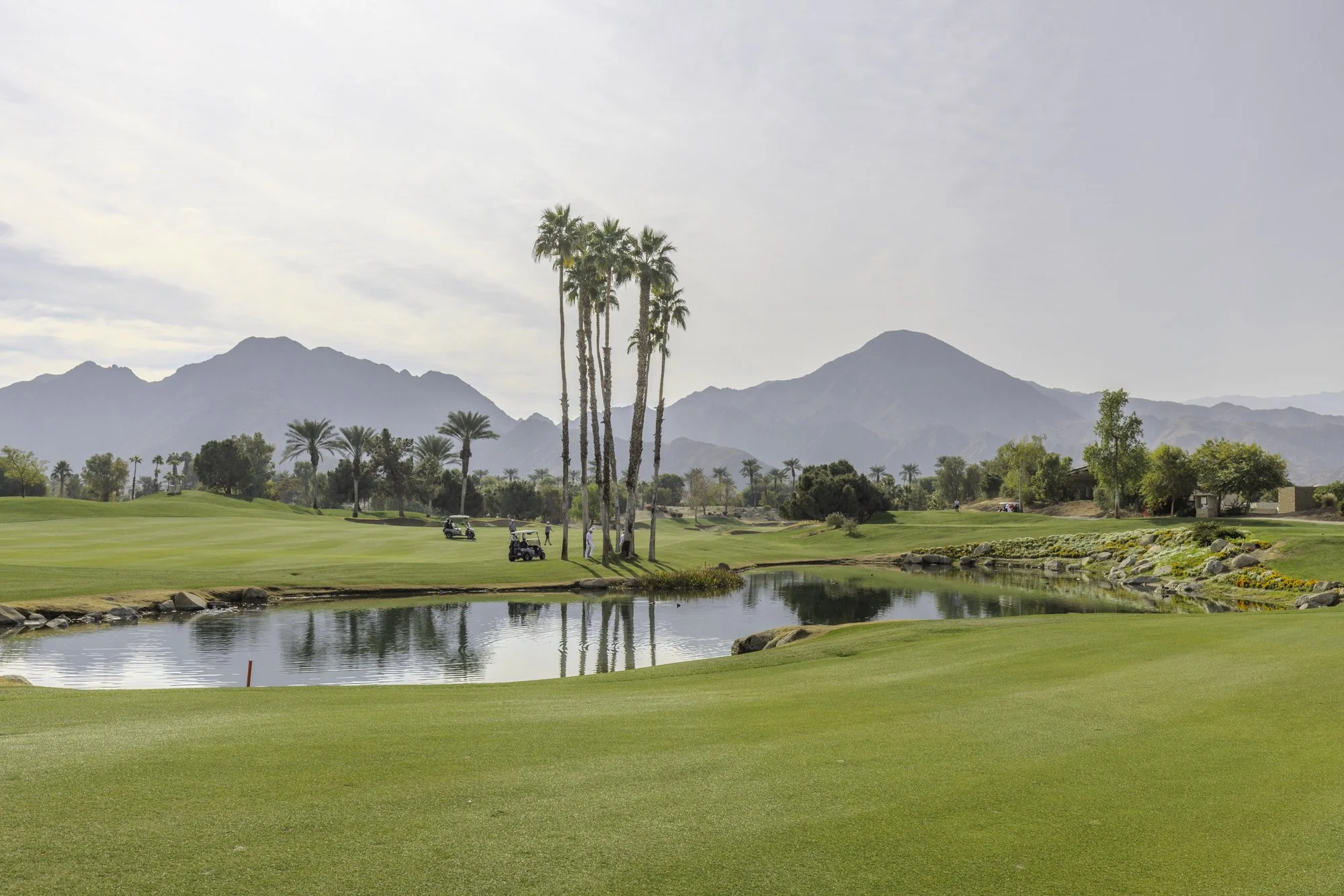 Golf course landscape with palm trees, water hazard, mountains in the background, and a cloudy sky.