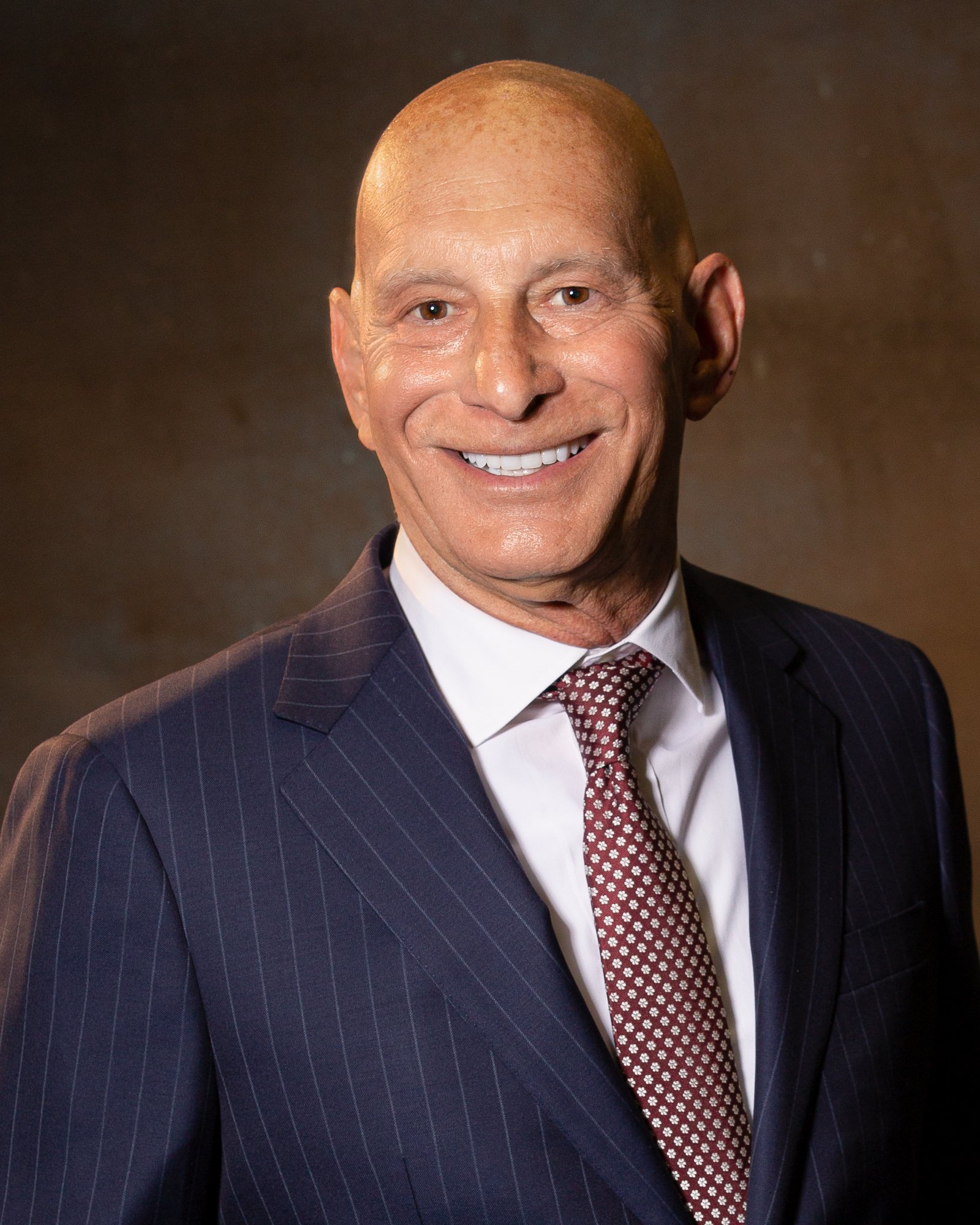 Bald man in a dark pinstripe suit with a white shirt and patterned tie, smiling against a brown backdrop.