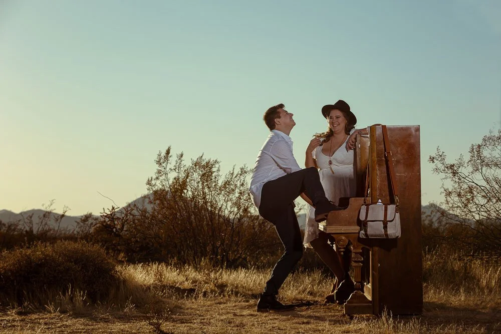Two people outdoors with a piano in a desert setting, one is standing on the piano bench and the other is leaning against the piano, surrounded by dry vegetation.