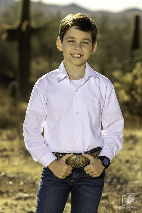 Young boy in a white shirt and jeans standing outdoors in a desert setting with cacti in the background.