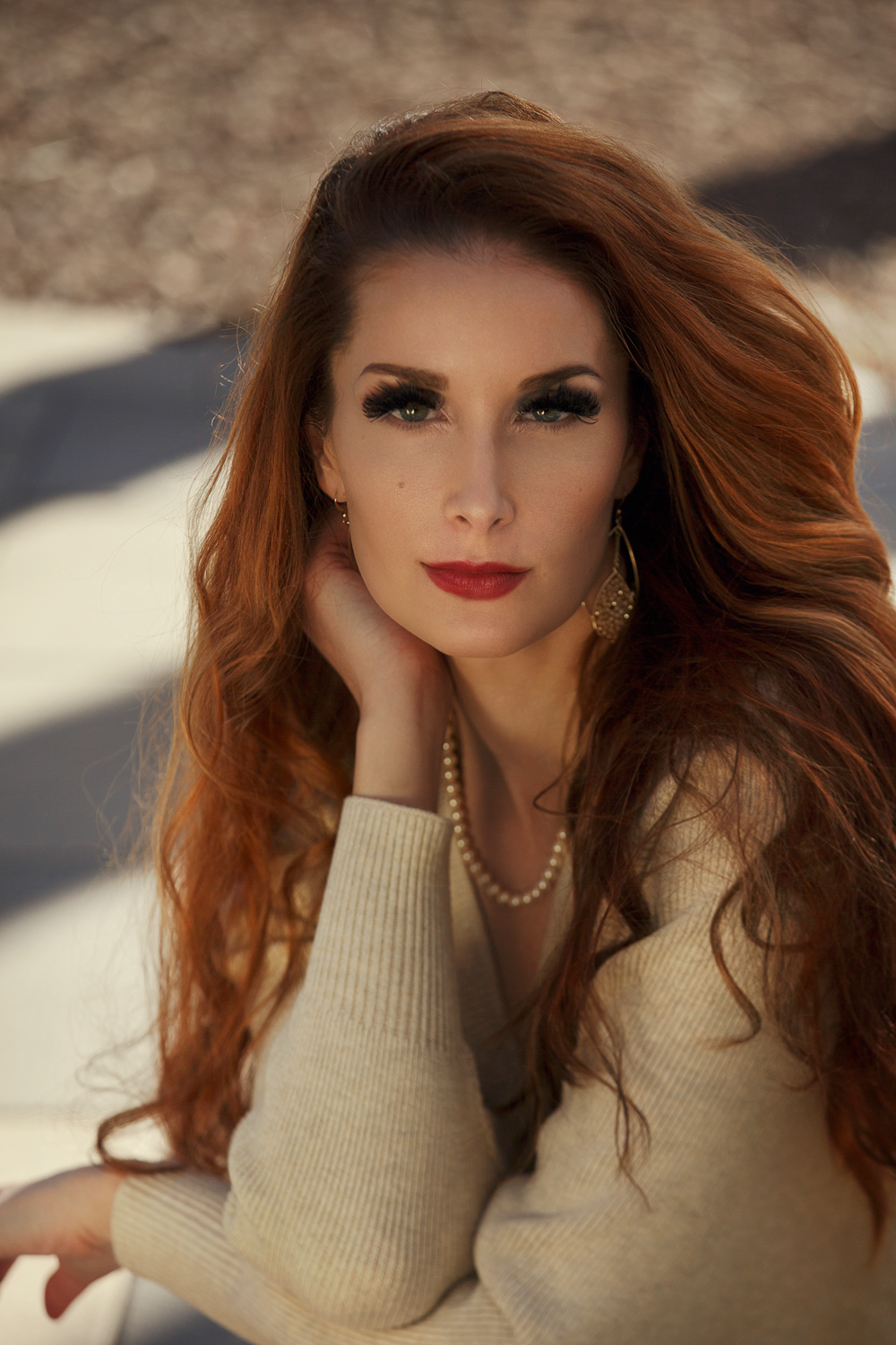 Woman with red hair and beige sweater, wearing pearl necklace, posed outdoors.