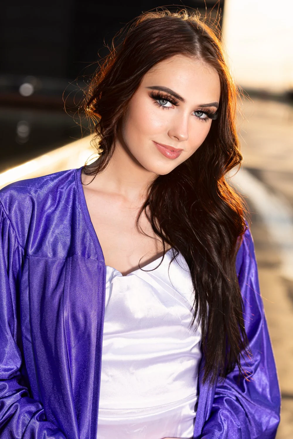 Woman with long dark hair wearing a purple graduation gown and a white top, outdoors with the sun setting in the background.